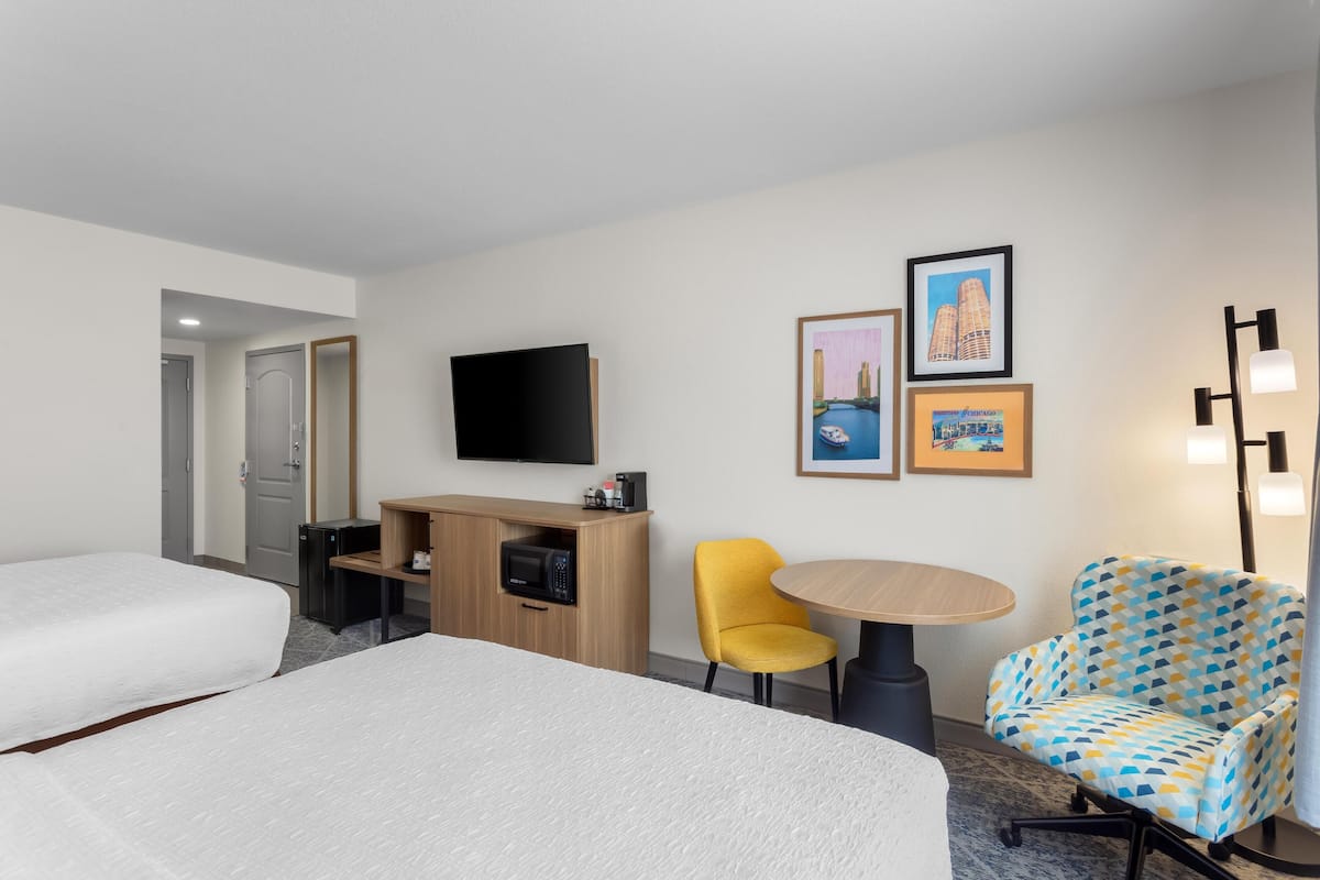 A hotel room with two beds, a television, a desk with a microwave, a yellow chair, a patterned armchair, and framed pictures on the wall.