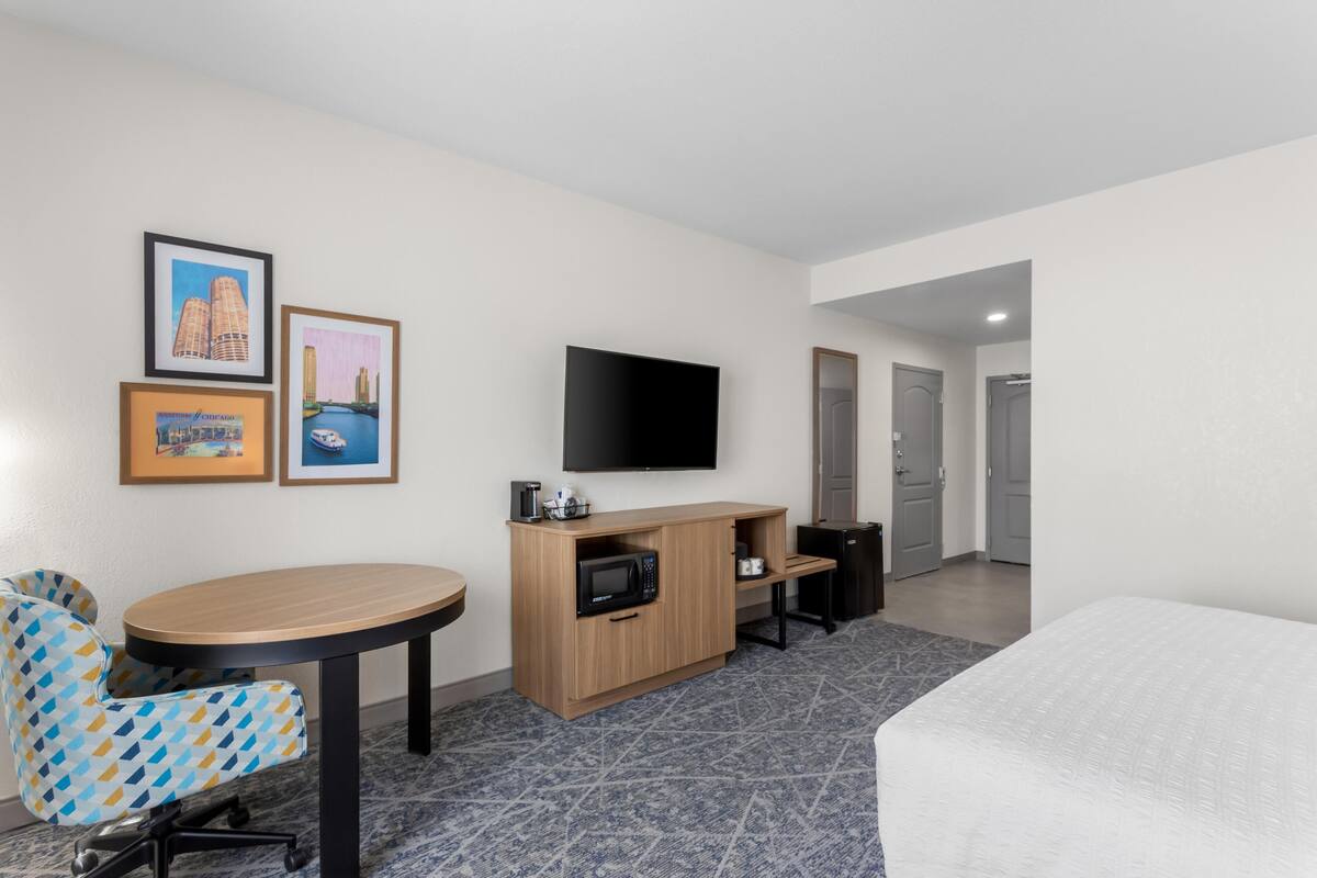 A hotel room with a bed, a desk with a flat-screen TV, a microwave, a coffee maker, a chair, and framed pictures on the wall.