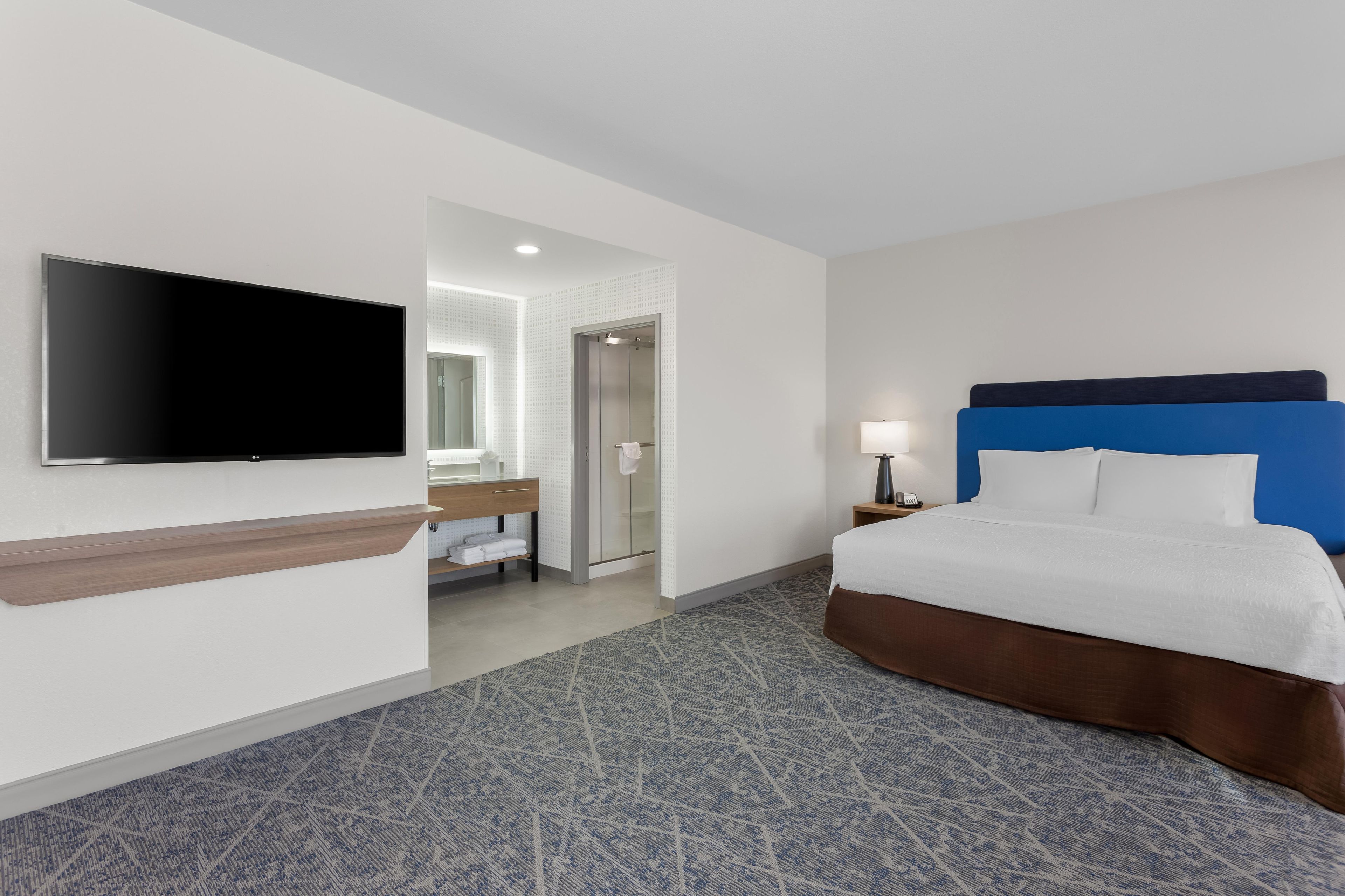 Habitación de hotel con cama, televisor montado en la pared, baño con espejo y mesita de noche con lámpara.