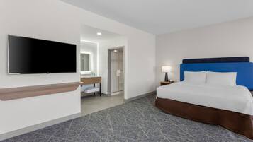 Habitación de hotel con cama, televisor montado en la pared, baño con espejo y mesita de noche con lámpara.