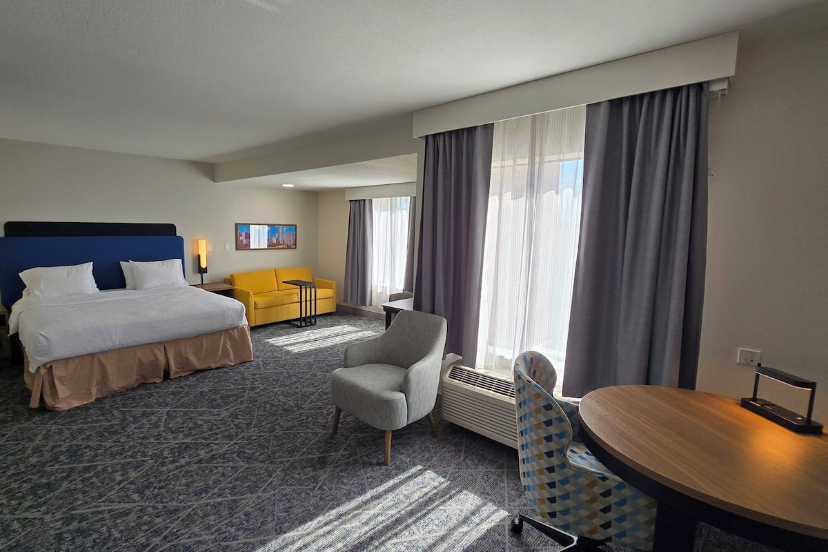 A hotel room with a bed, a desk, a chair, a sofa, and a large window with curtains.