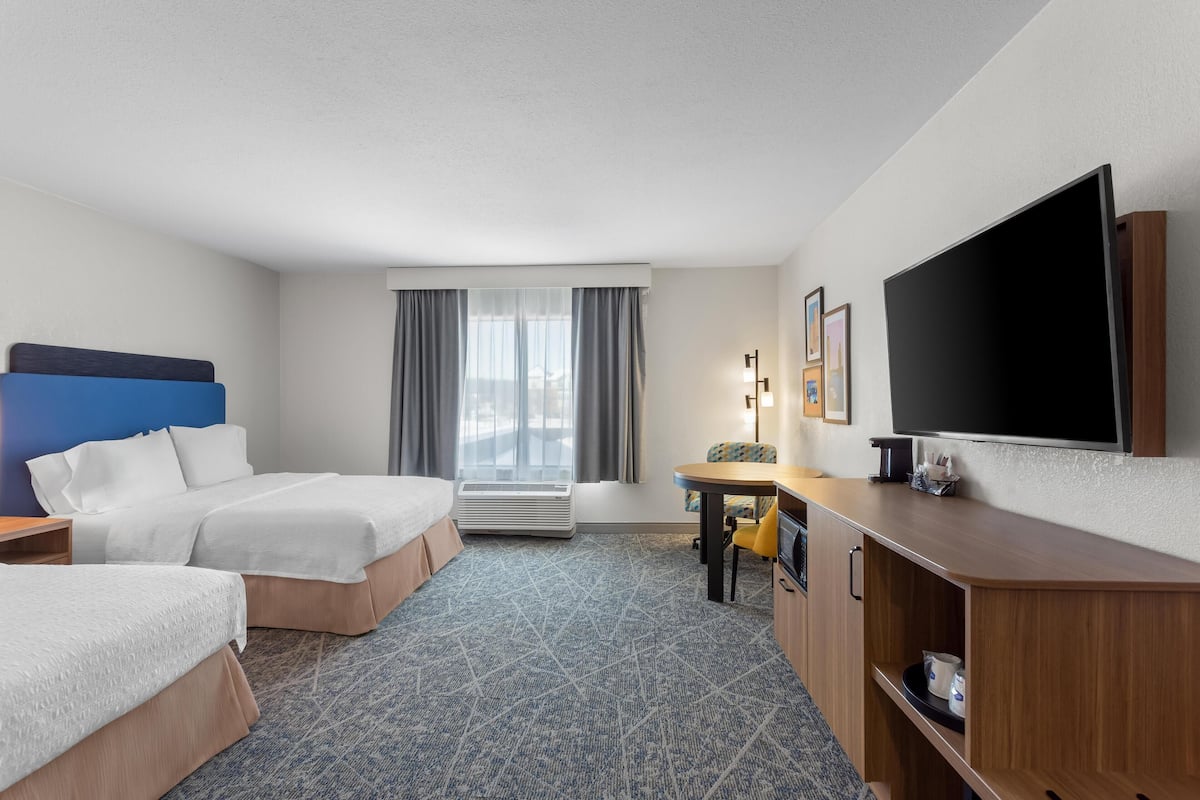 A hotel room with two beds, a desk, a flat-screen TV, a lamp, and a window with curtains.