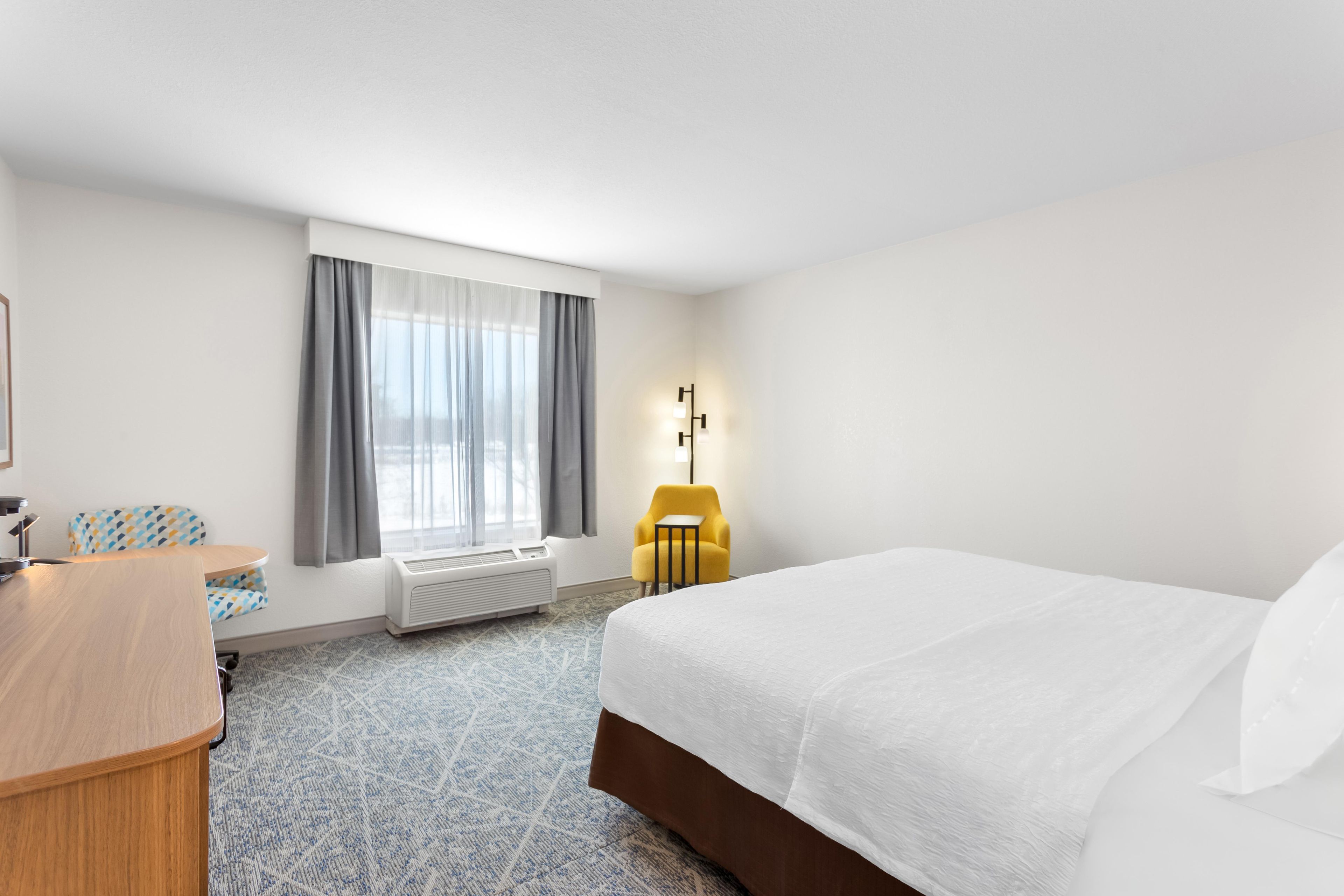A hotel room with a bed, a desk, a chair, a radiator, and a window with curtains.