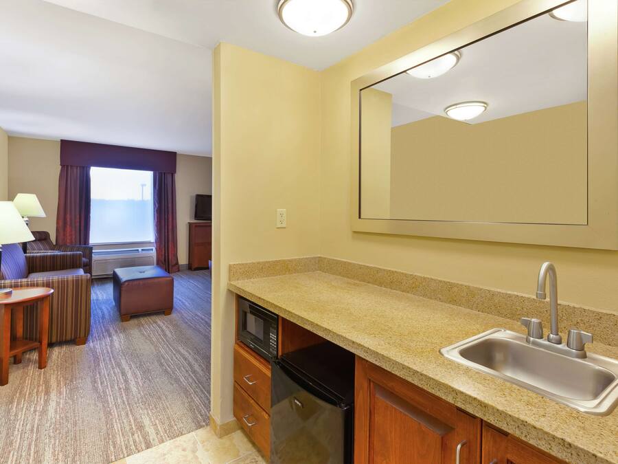 A compact hotel room with a kitchenette, a large mirror, a sofa, and a window with curtains.