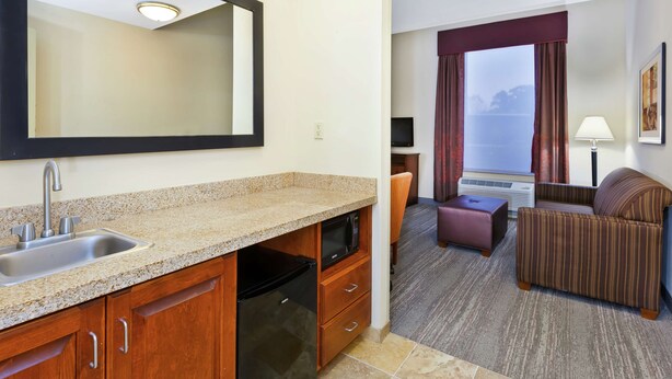 Una habitación de hotel con kitchenette, zona de estar con sofá y sillón, y un ventanal con cortinas.