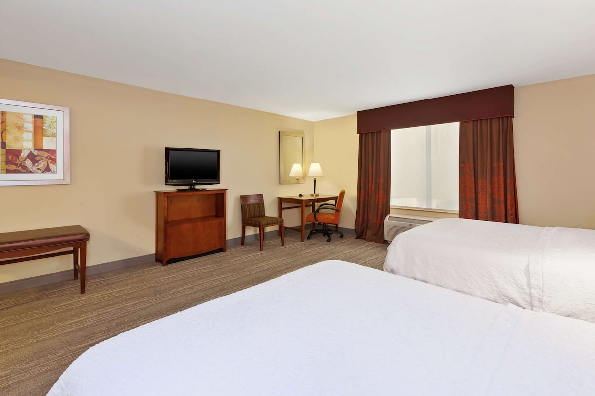 A hotel room with two beds, a desk with a chair, a television, a mirror, and a window with curtains.