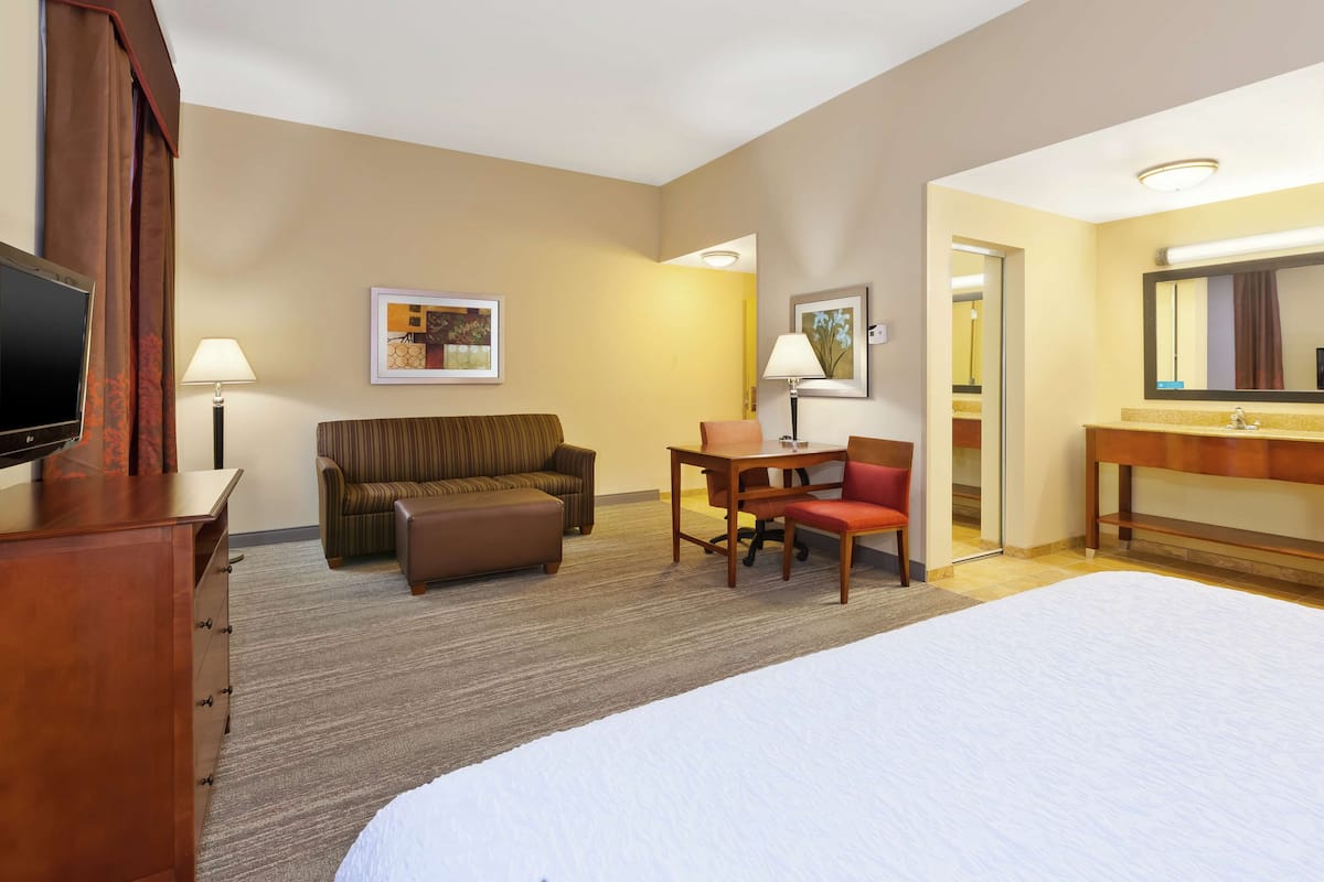 A hotel room with a bed, a desk with a chair, a sofa, a TV, and a bathroom mirror.