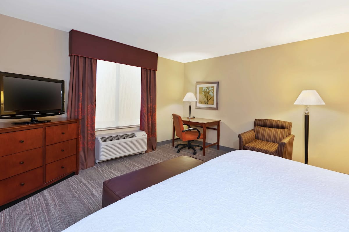 A hotel room with a bed, a desk with a chair, a television, an air conditioner, and two lamps.