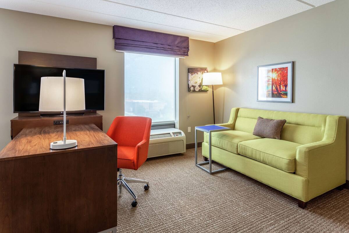 A hotel room with a wooden desk, a television, a red chair, a green sofa, a small table, a lamp, and a window with a view.