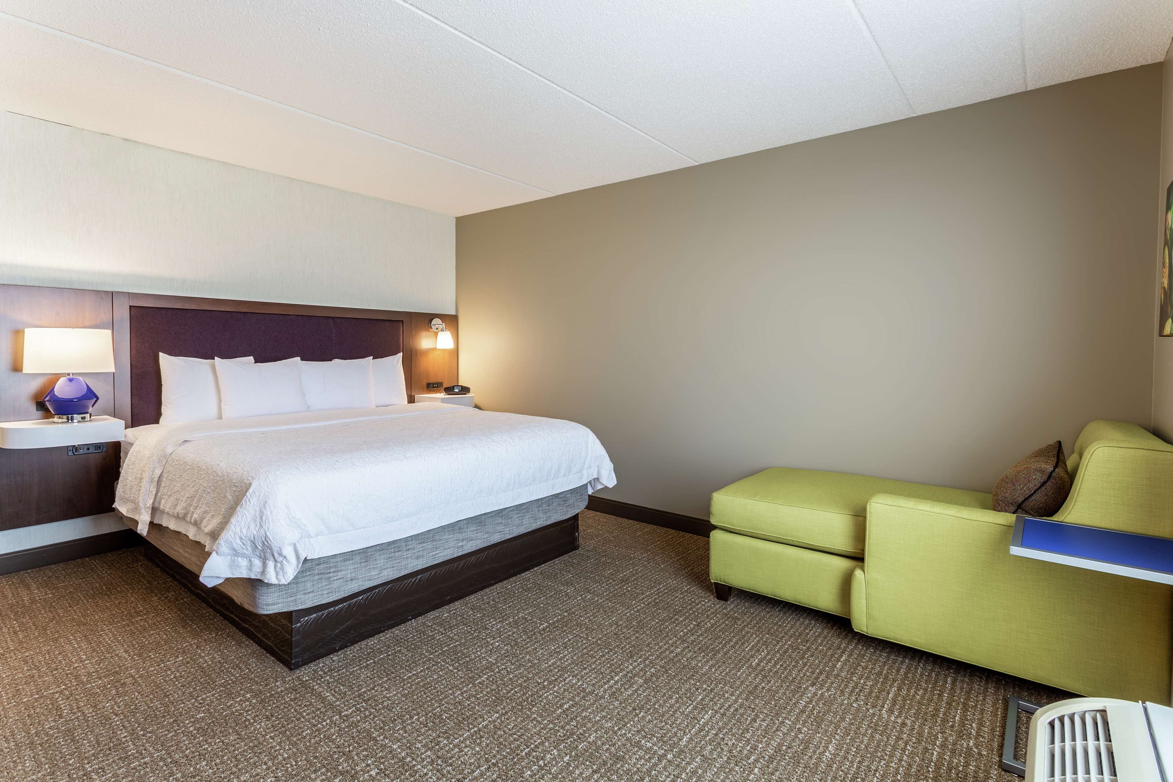 Une chambre d’hôtel avec un lit, une table de chevet, une lampe, un fauteuil vert et une petite table.