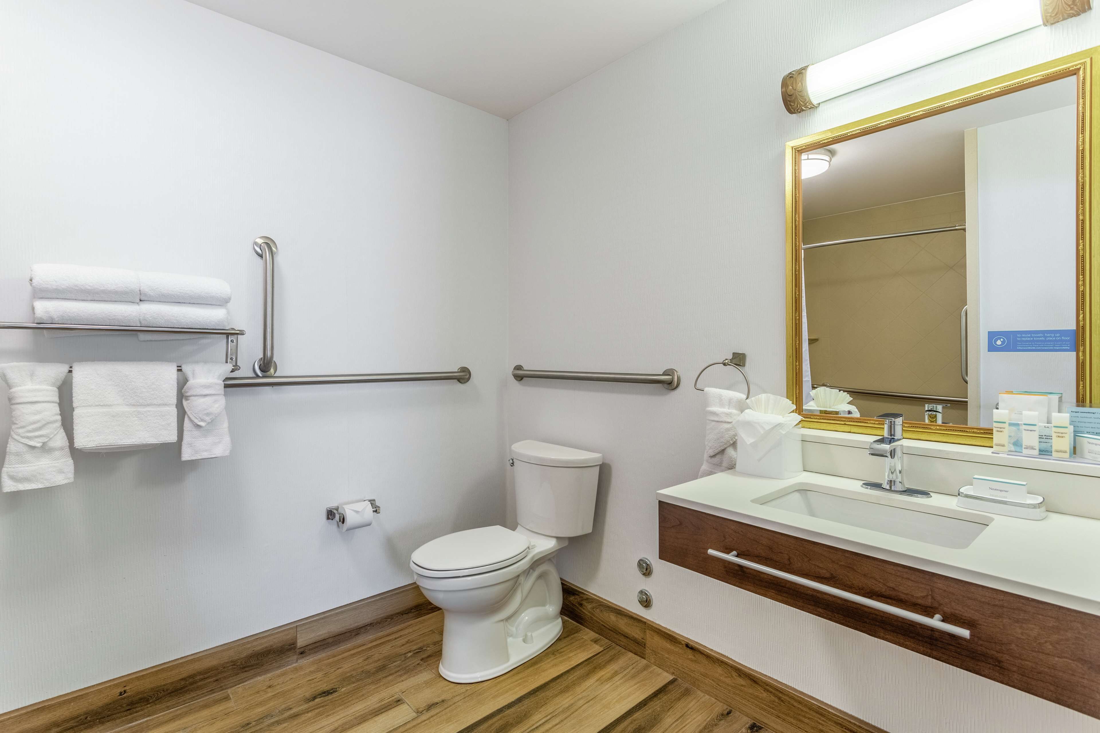 Un baño con inodoro, barras de apoyo, lavamanos y espejo.