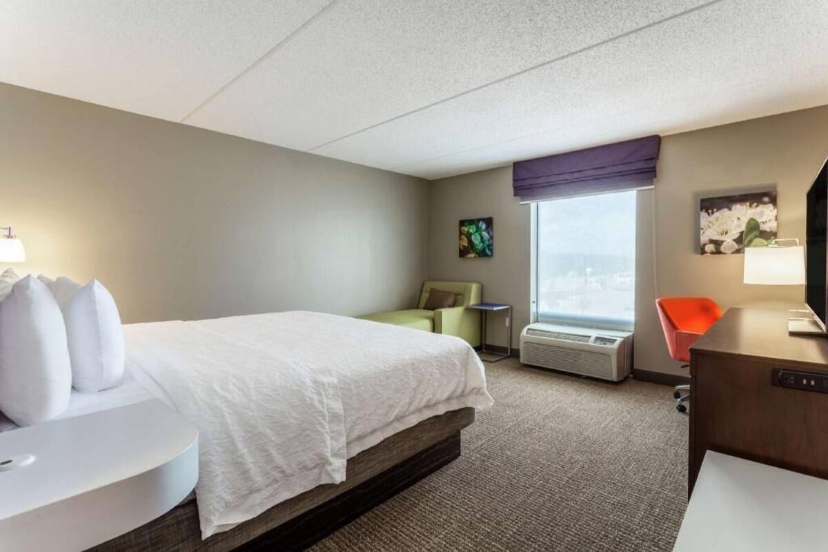 A hotel room with a bed, a desk, a chair, a window with a view, and an air conditioning unit.