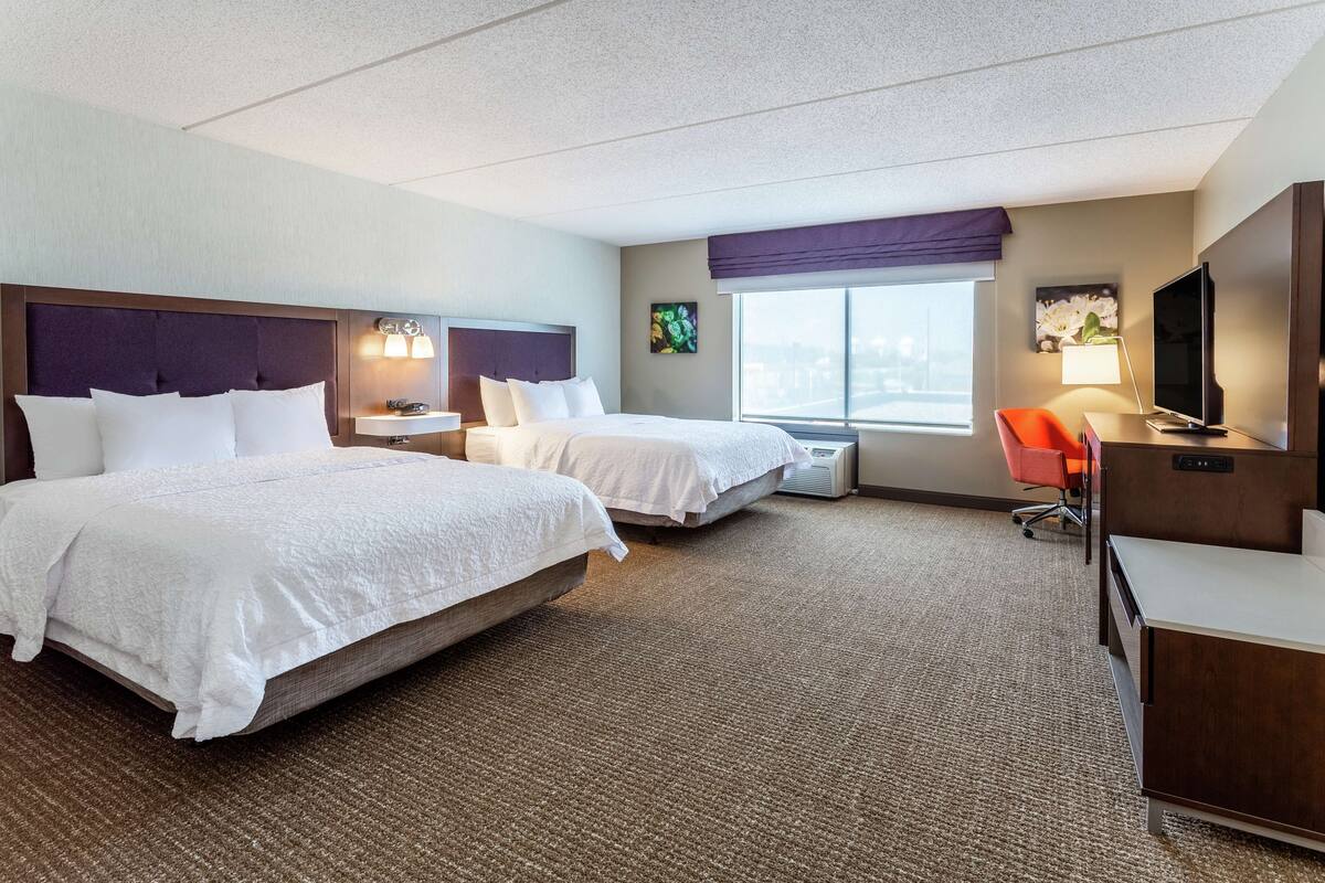 A hotel room with two beds, a desk, a TV, and a window with a city view.