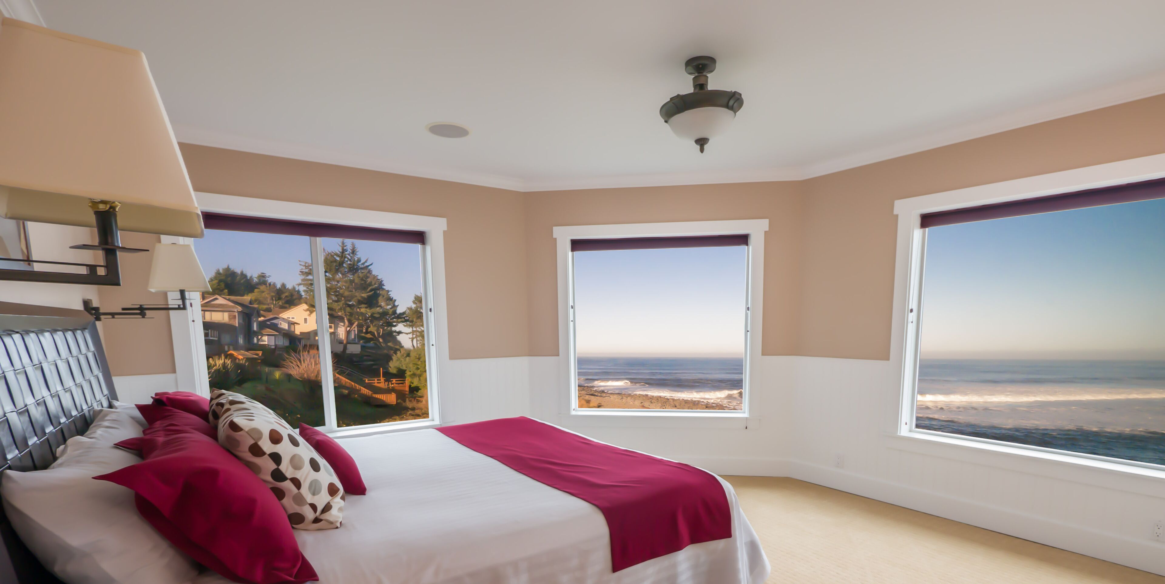 A bedroom with a bed, two windows with ocean views, a ceiling fan, and a nightstand with a lamp.