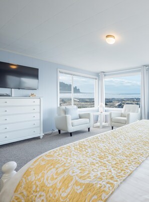 A modern bedroom with a large bed, a TV, a dresser, two chairs, and a view of the ocean. - Seaside Vacation Homes (Seaside)