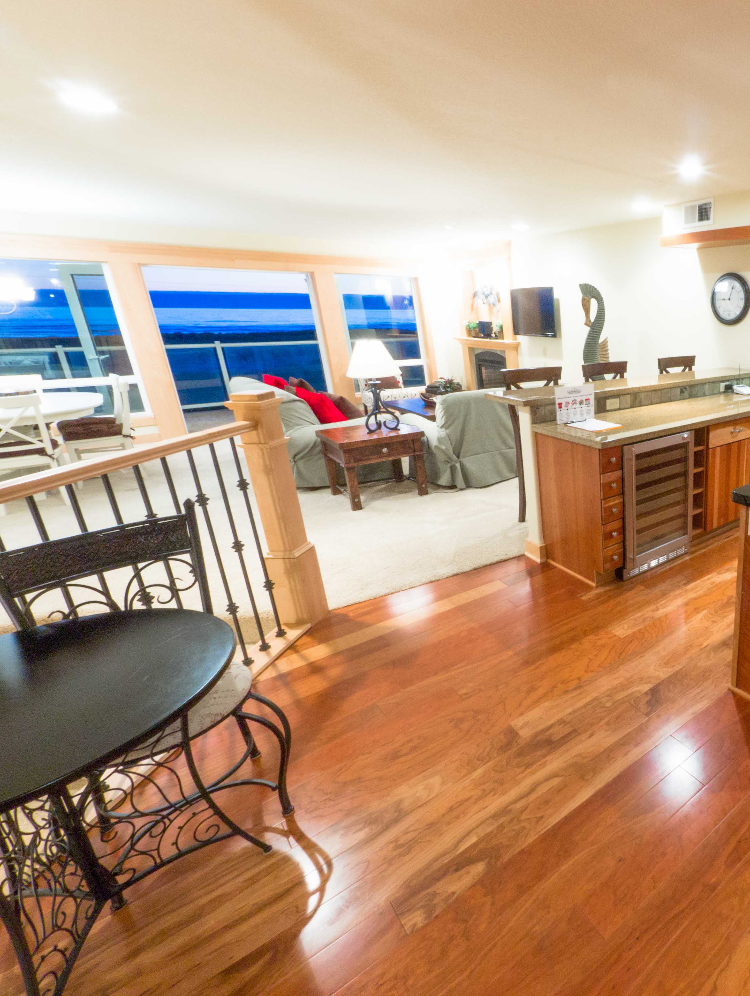 A spacious living area with a wooden floor, a dining table with chairs, a sofa, and a TV. There are large windows with a view of the ocean.