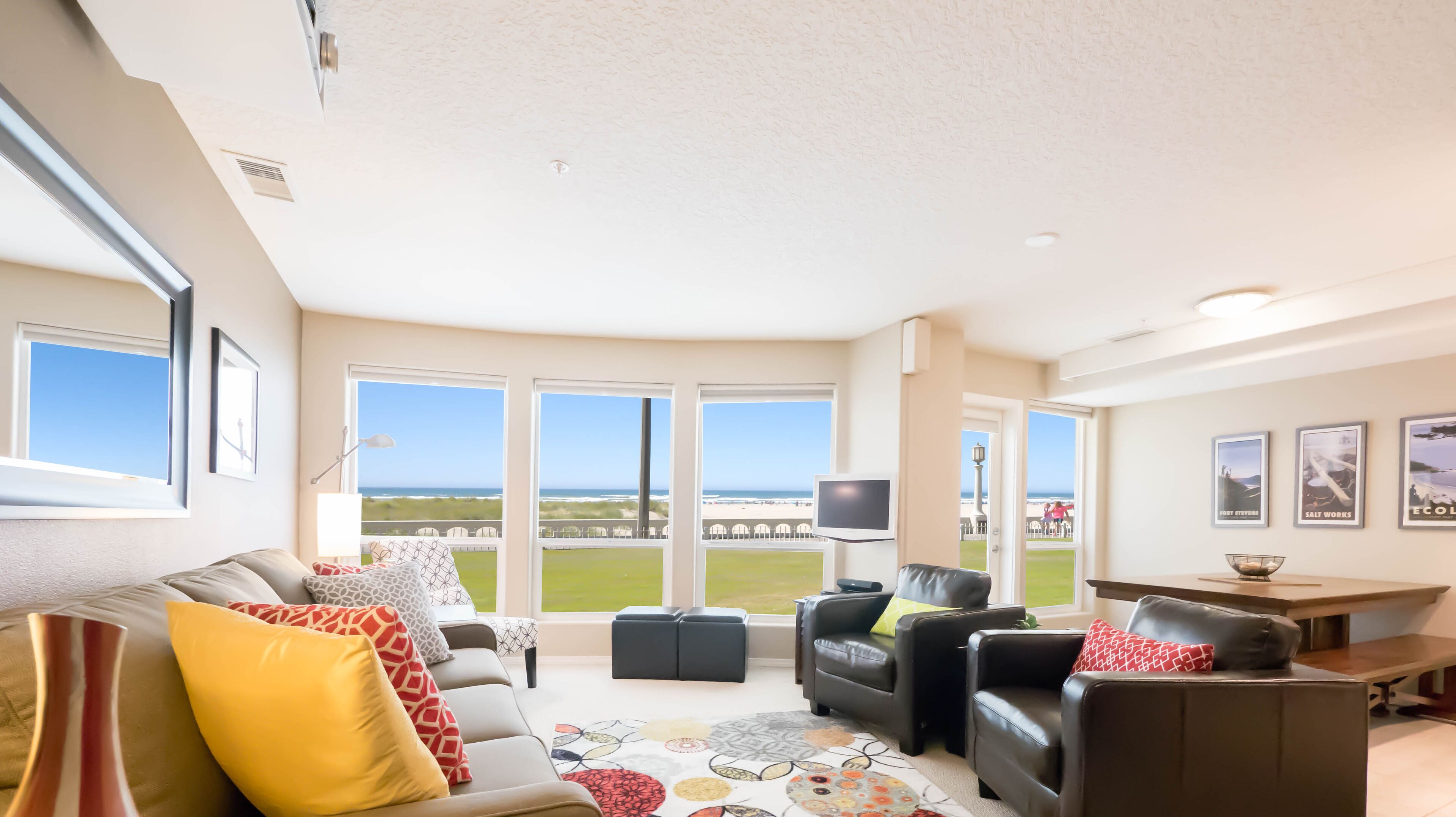 A spacious living room with a large window offering a view of the beach. There is a sofa, a coffee table, and a TV.