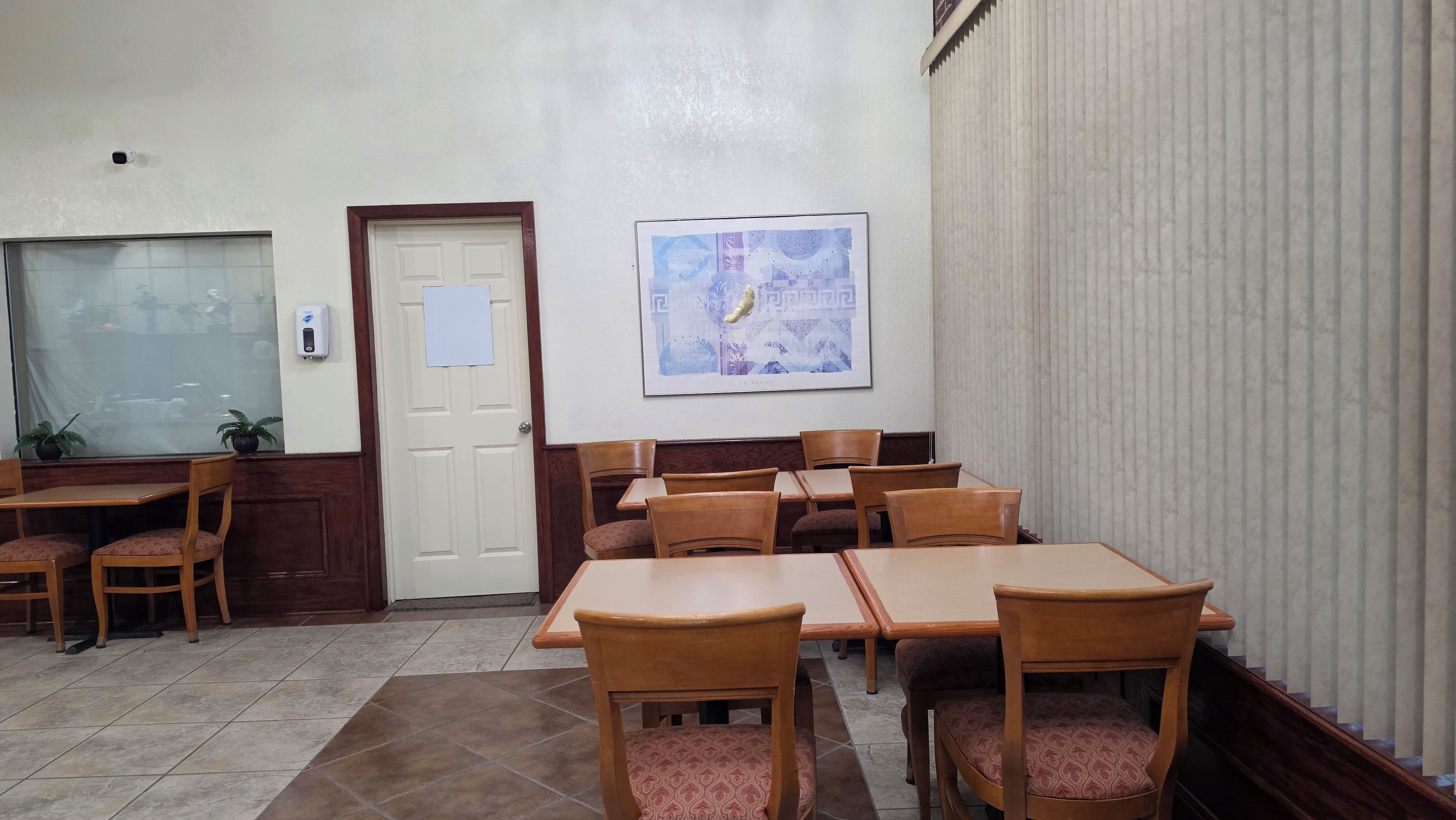 A hotel room with a large window, a door with a paper on it, a framed picture on the wall, and several wooden tables and chairs.