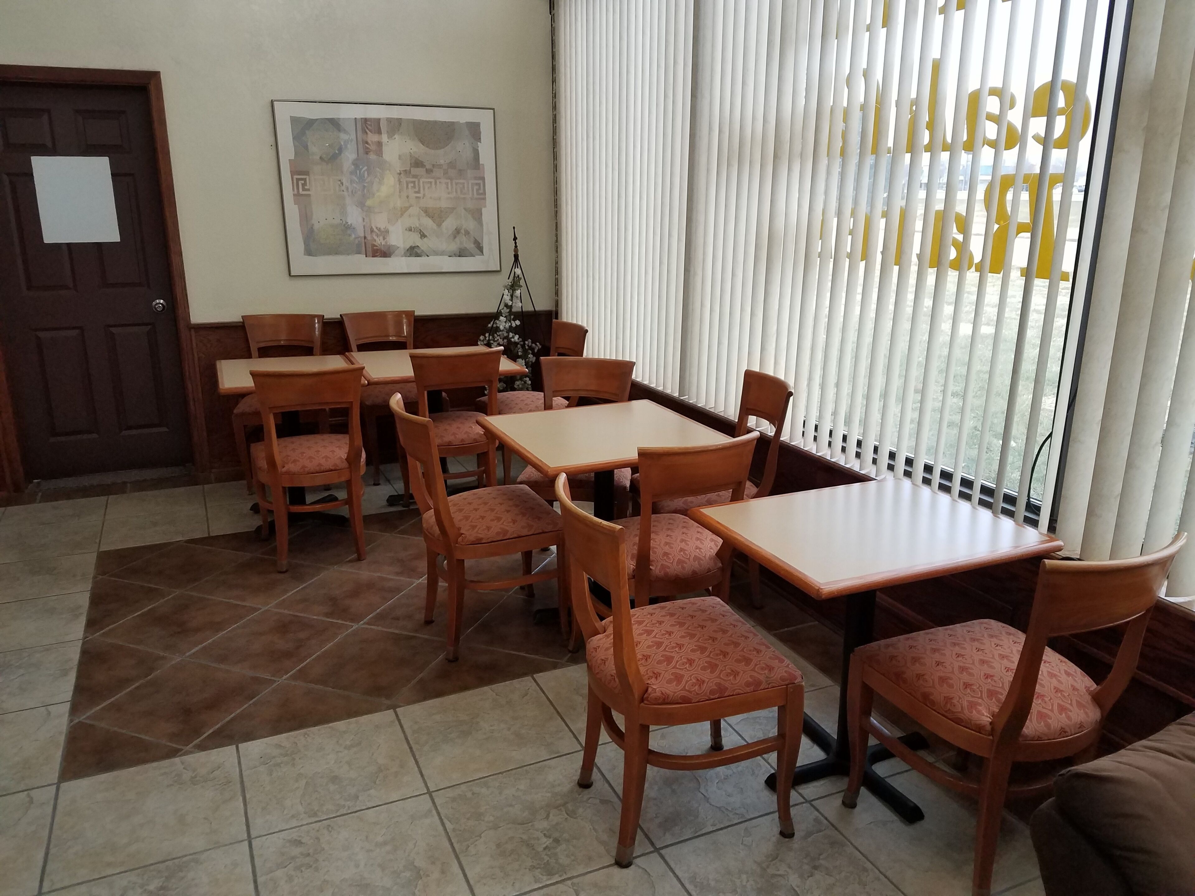 A dining area with wooden tables and chairs, a door, a framed artwork, and a window with blinds.