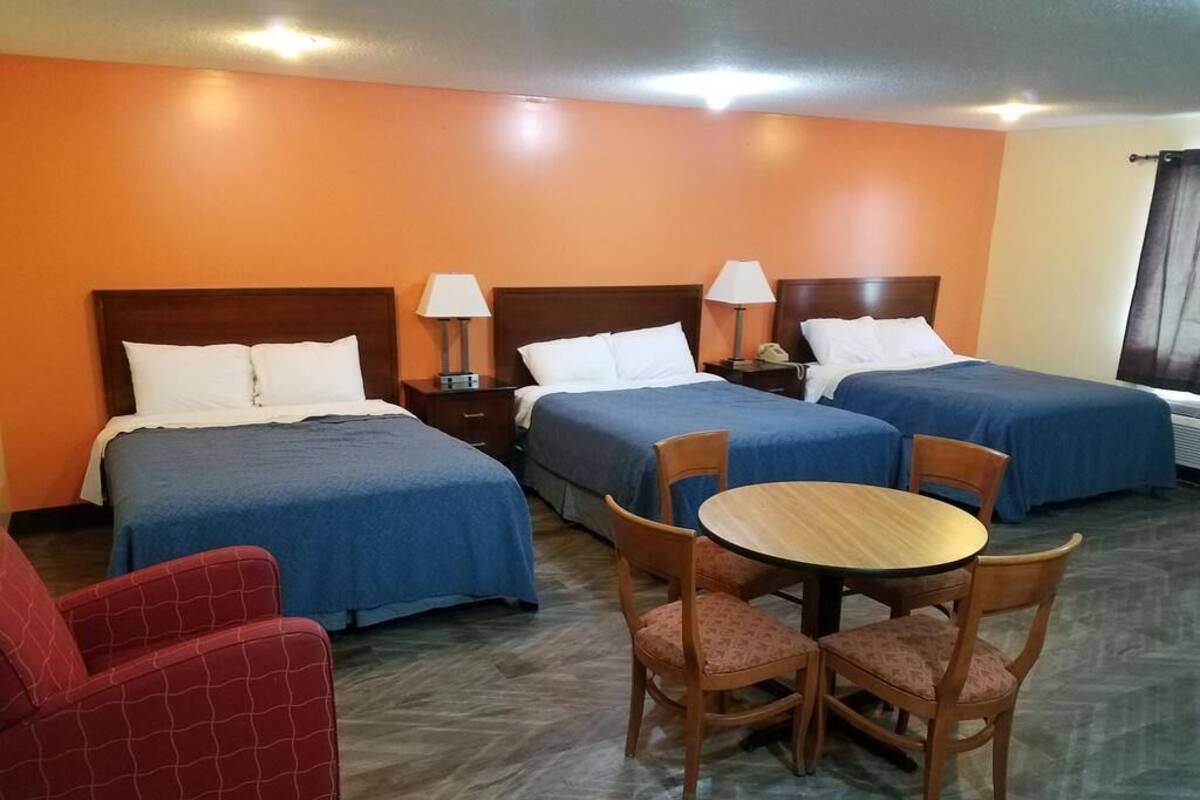A hotel room with three beds, a round wooden table with chairs, and a red armchair.