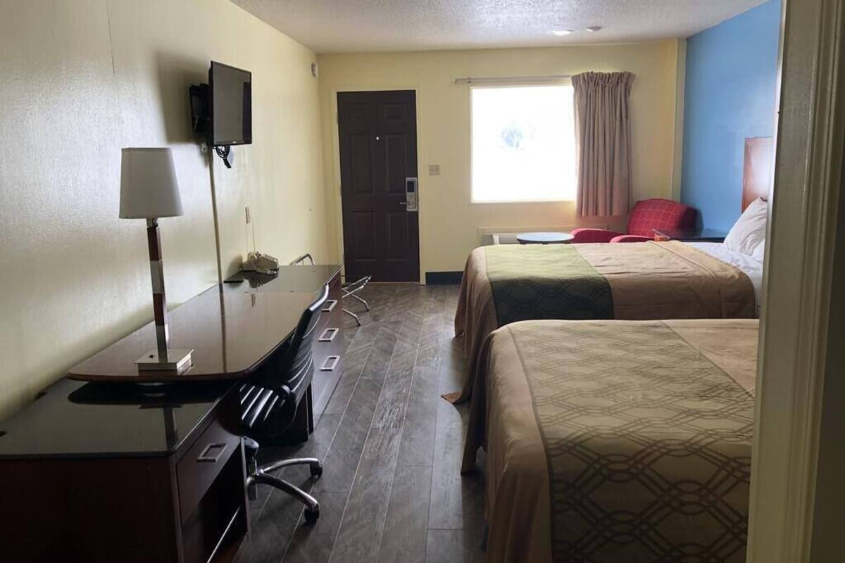 A hotel room with a desk, two beds, a chair, a TV, and a window with curtains.