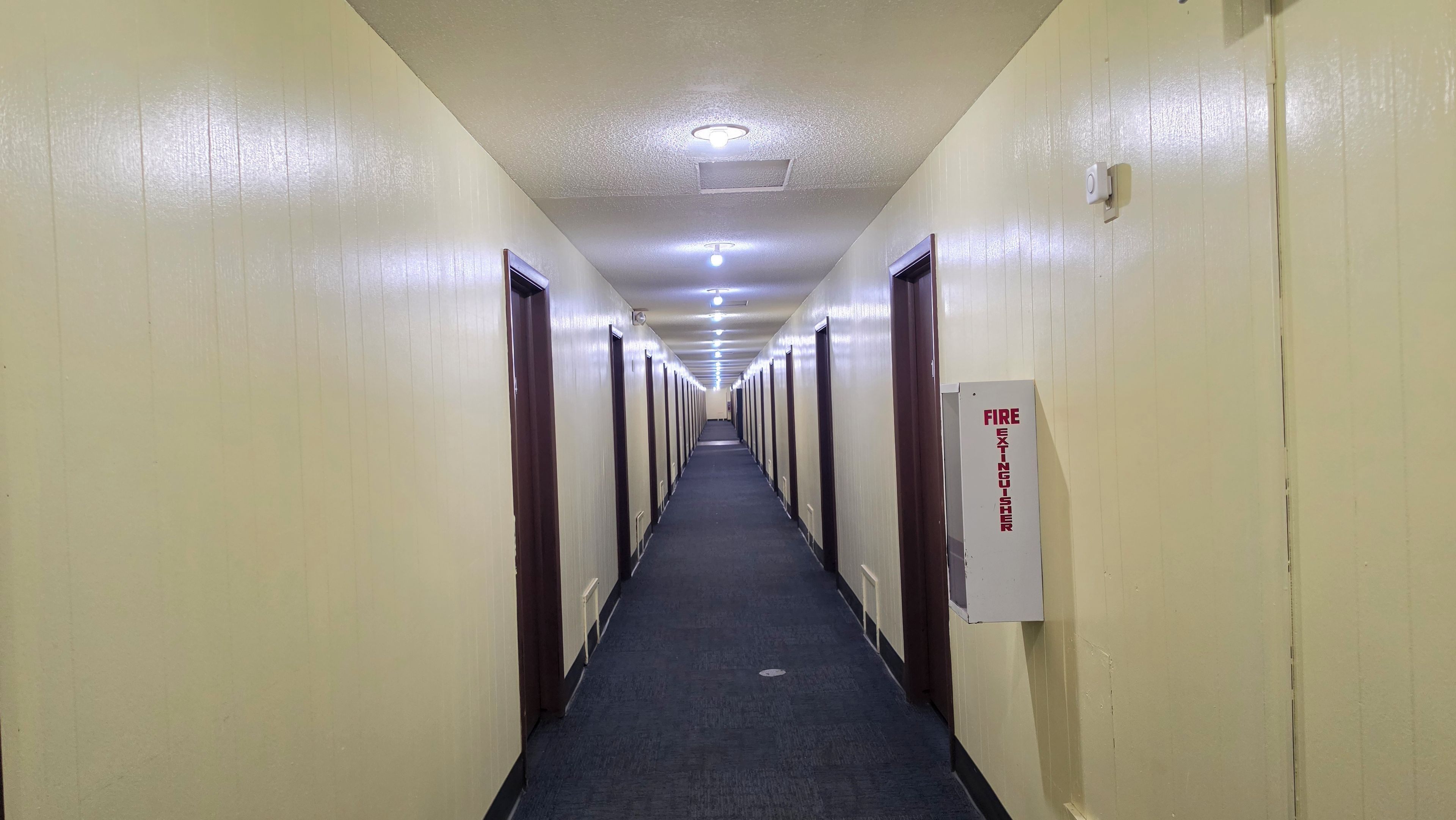 Un pasillo de hotel con puertas a ambos lados, un extintor en la pared derecha y una lámpara en el techo.