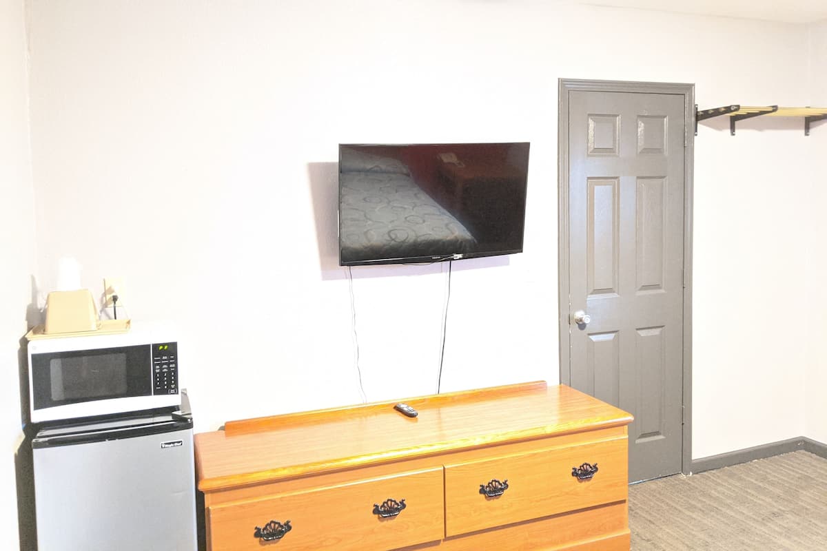 A compact room with a flat-screen TV mounted on the wall, a microwave, and a wooden dresser with drawers.