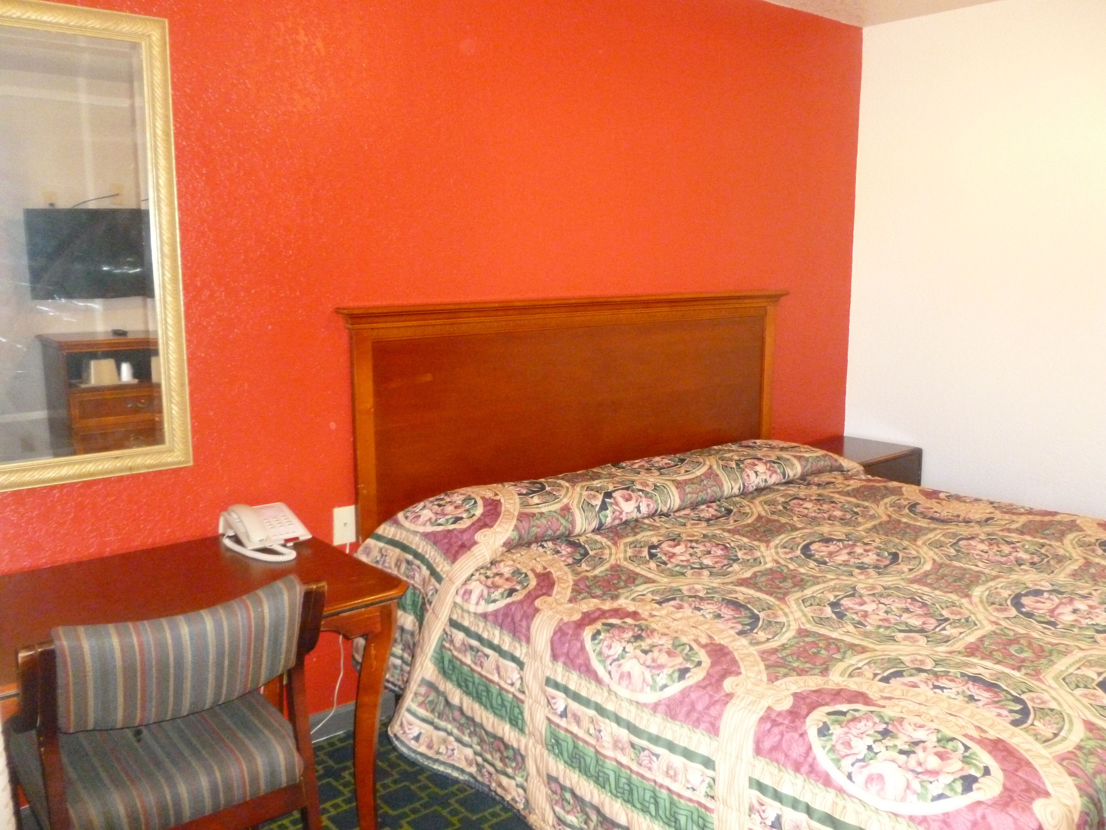 Habitación de hotel con cabecera de madera, cama con colcha de motivos florales, una pequeña mesa de trabajo con teléfono y una silla.