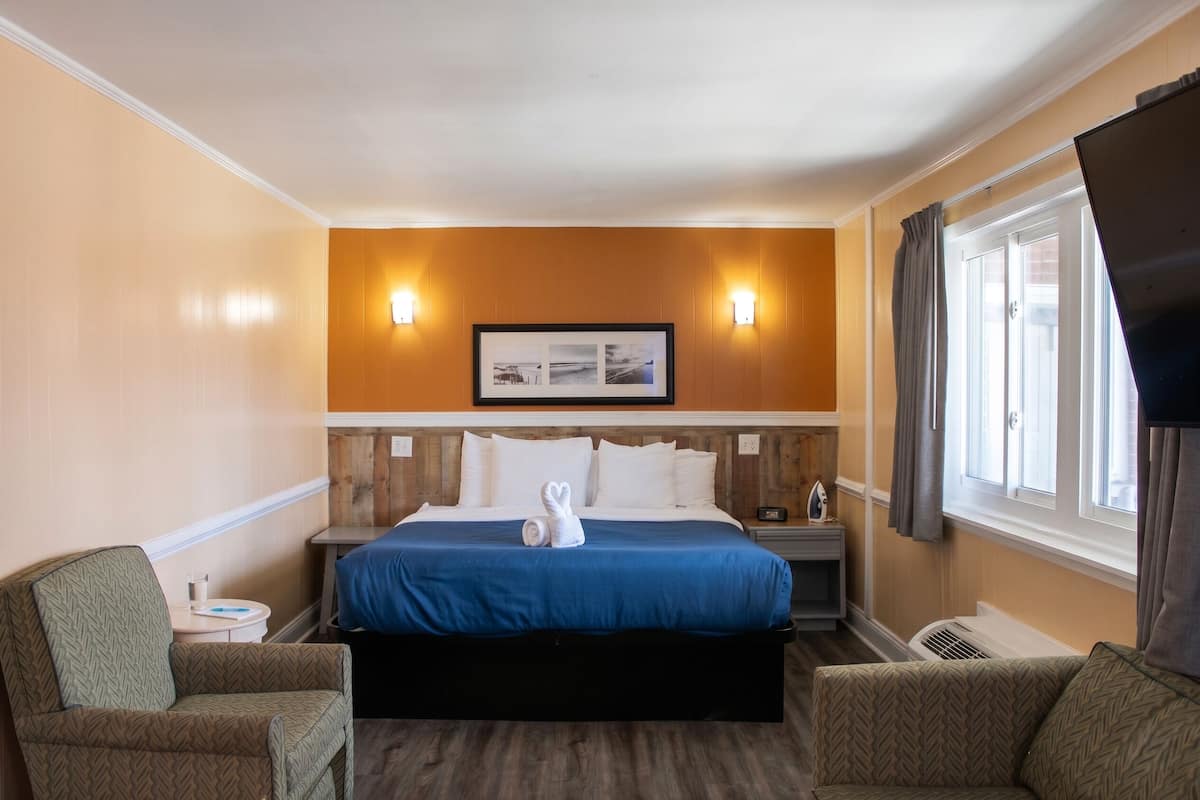 A hotel room with a bed, two chairs, a nightstand, a picture frame, and a window with curtains.