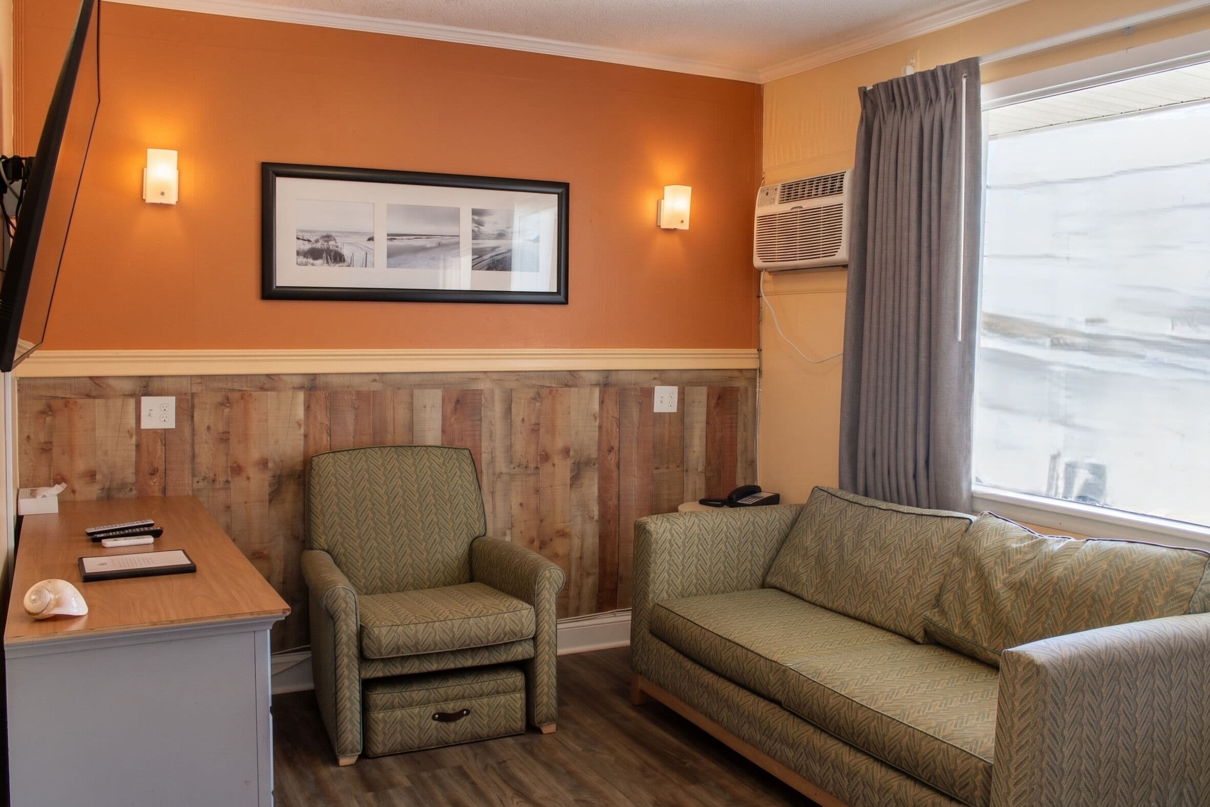 Une chambre d’hôtel avec un mur lambrissé, un canapé, un fauteuil, un bureau avec un clavier et des télécommandes, et un tableau encadré accroché au mur.
