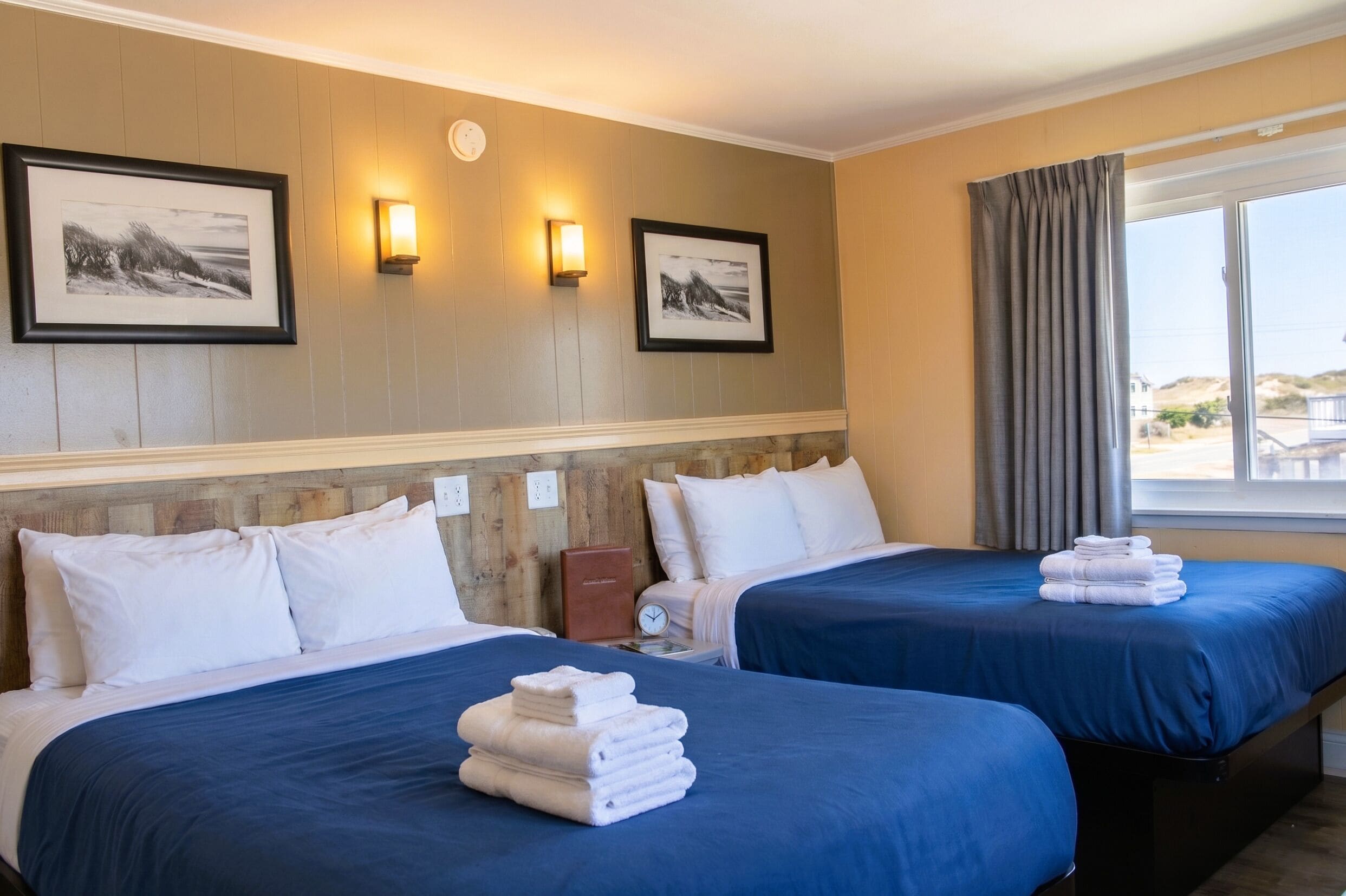 A hotel room with two beds, a wooden headboard, wall-mounted lights, framed pictures, a clock, and a window with curtains.