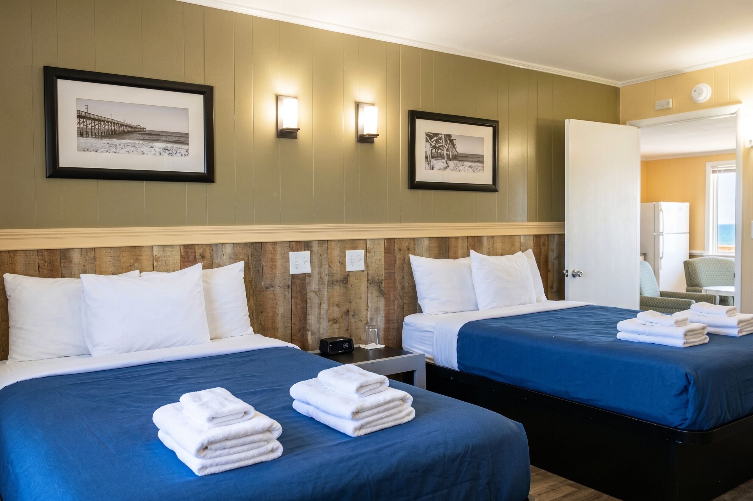 A hotel room with two beds, a wooden headboard, framed pictures on the wall, and a view of the beach through the open door.