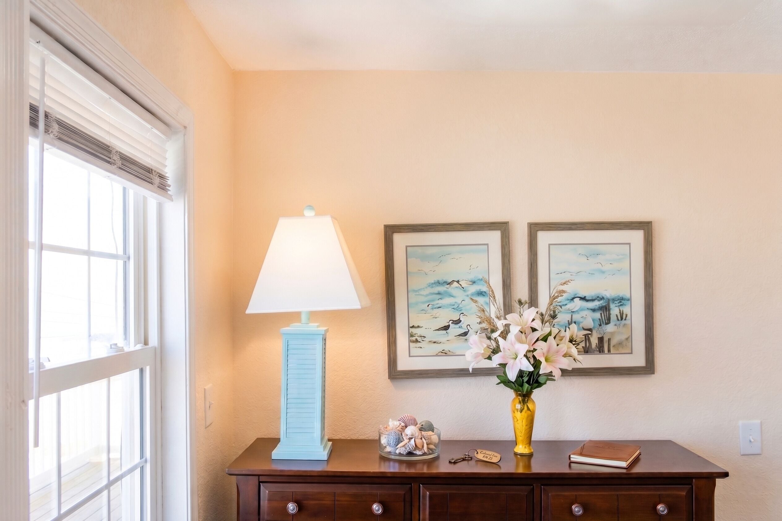 Une pièce avec une lampe, un vase de fleurs, deux tableaux encadrés et une commode en bois.