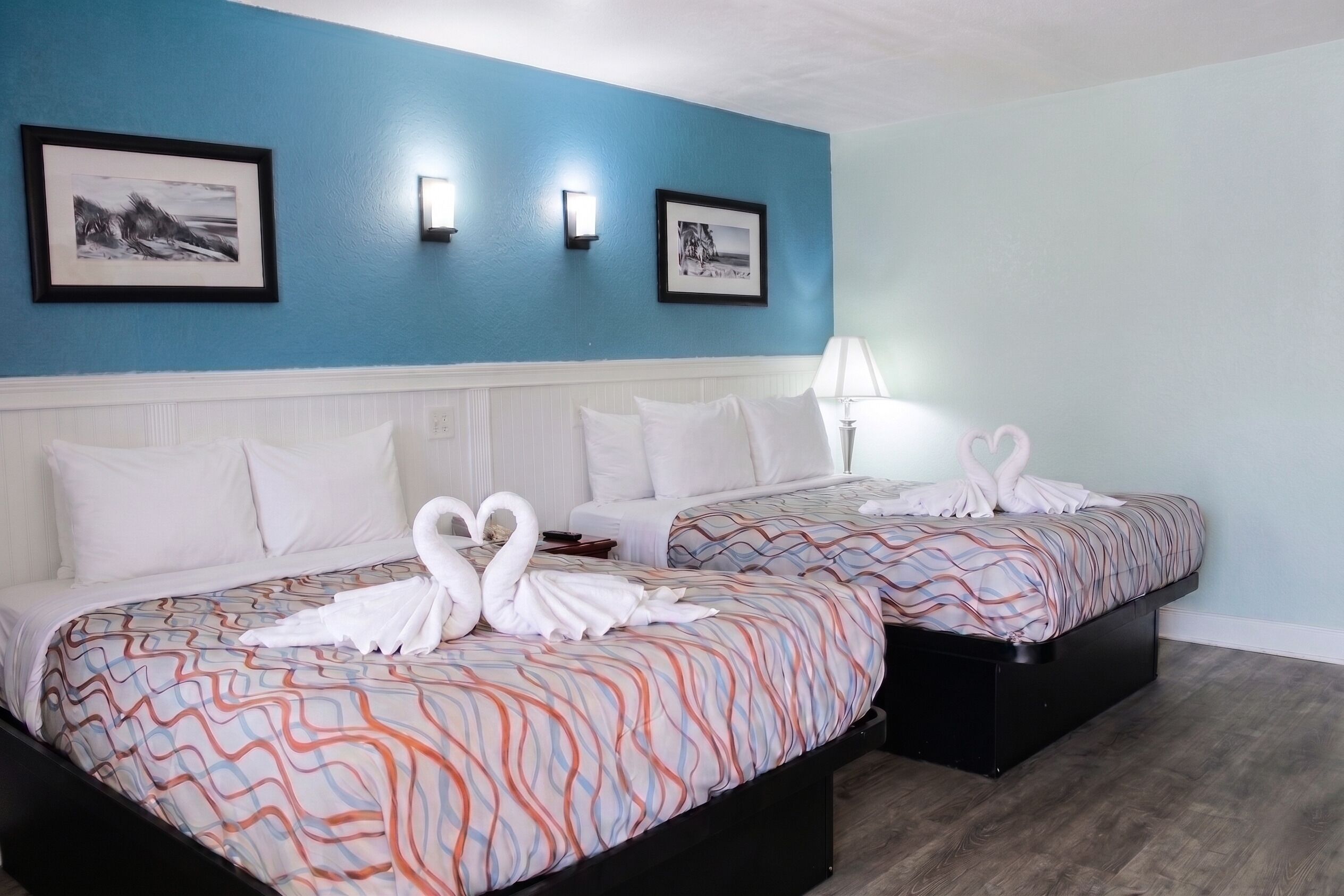 A hotel room with two beds, each with a patterned bedspread, white pillows, and a heart-shaped towel arrangement. There are two framed pictures on the wall and a bedside lamp.