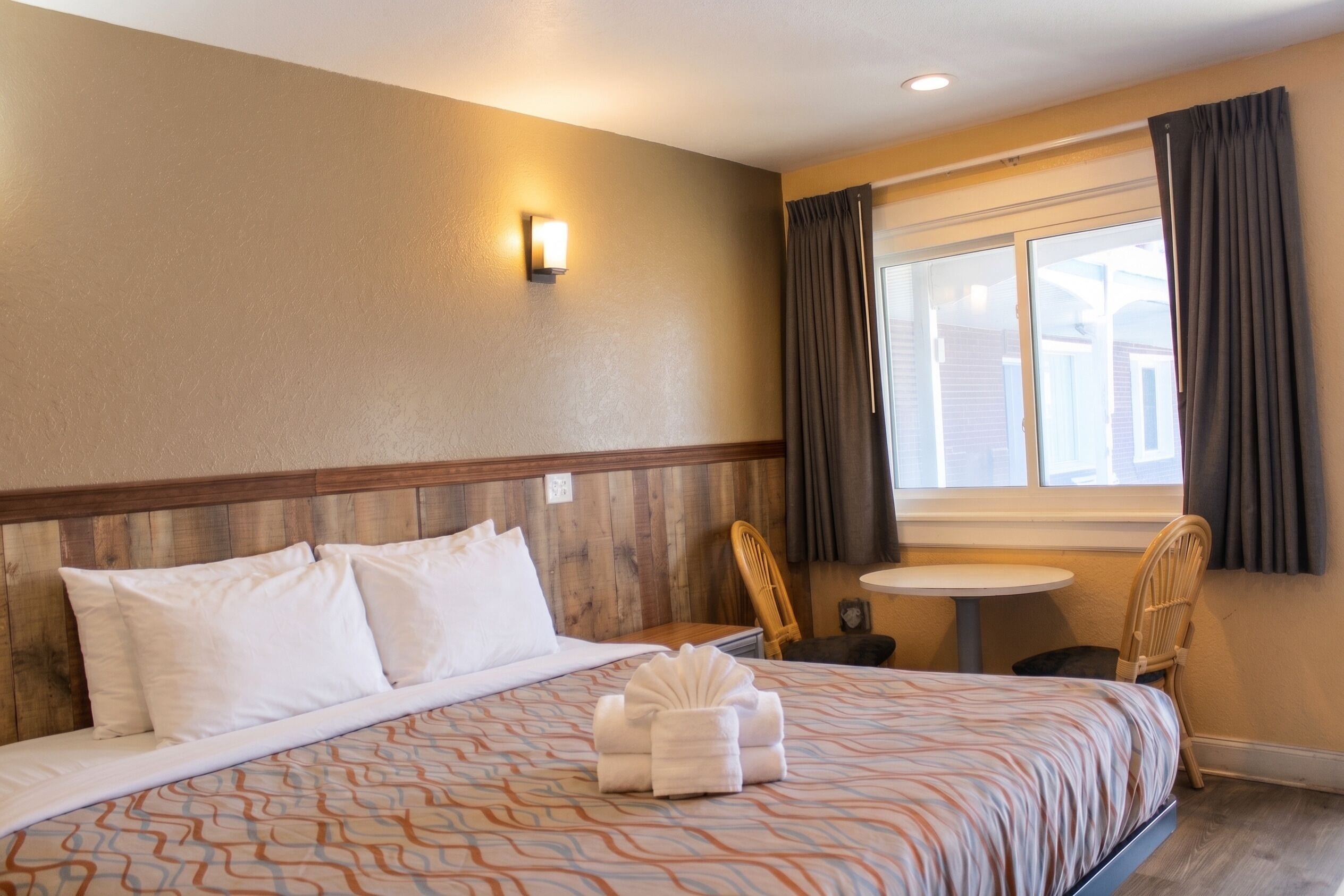 Habitación de hotel con cama, una mesita, una silla, ventana con cortinas y una lámpara fijada a la pared.