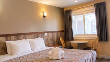 Habitación de hotel con cama, una mesita, una silla, ventana con cortinas y una lámpara fijada a la pared.