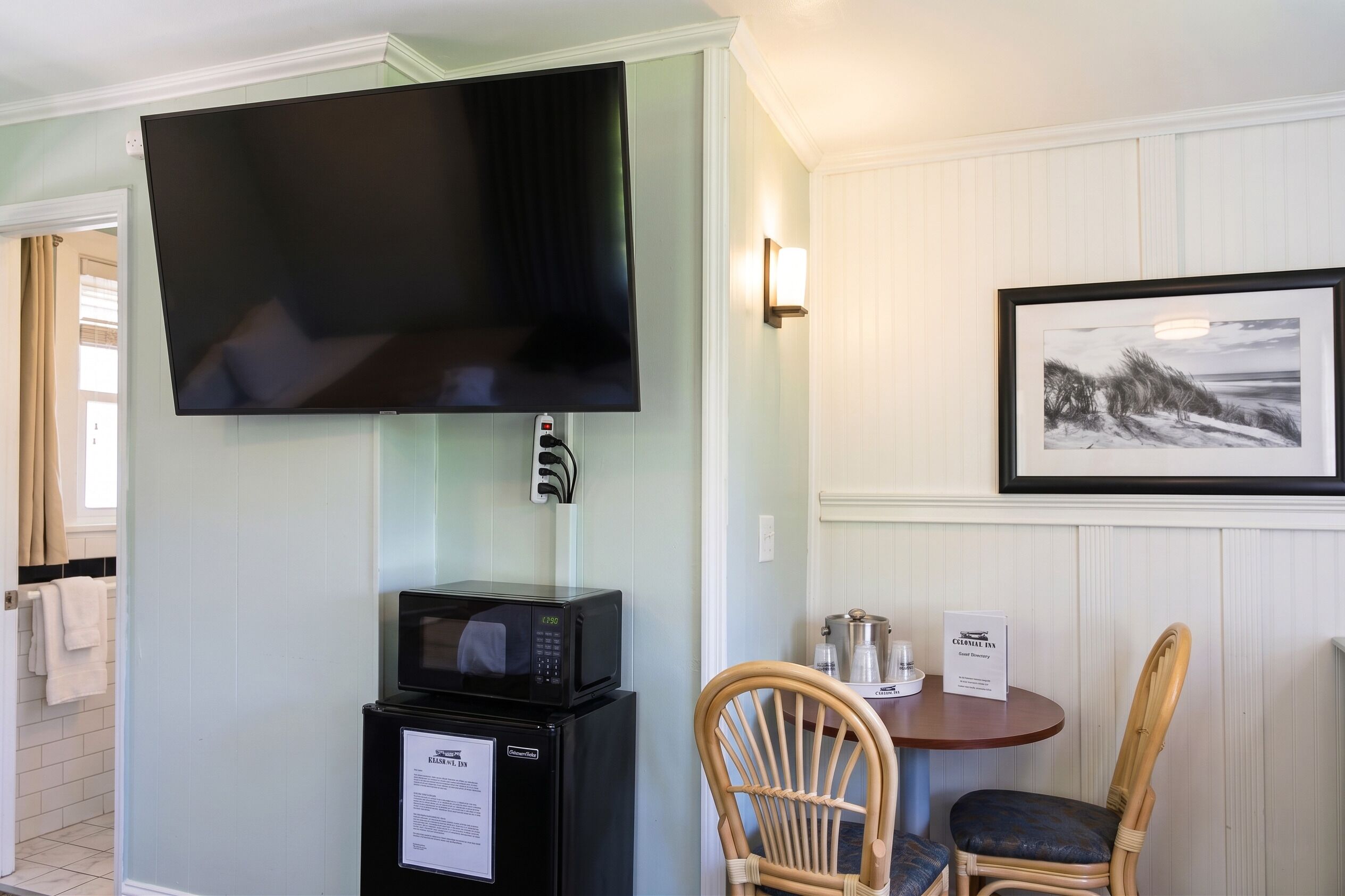 A room with a flat-screen TV mounted on the wall, a microwave, a refrigerator, a dining table with chairs, and a framed picture on the wall.