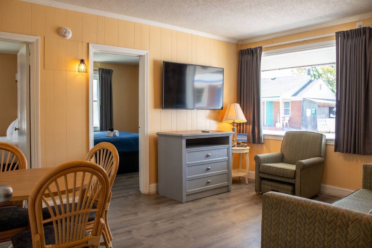 A room with a television, wooden dining table, chairs, a sofa, a lamp, and a window with curtains.