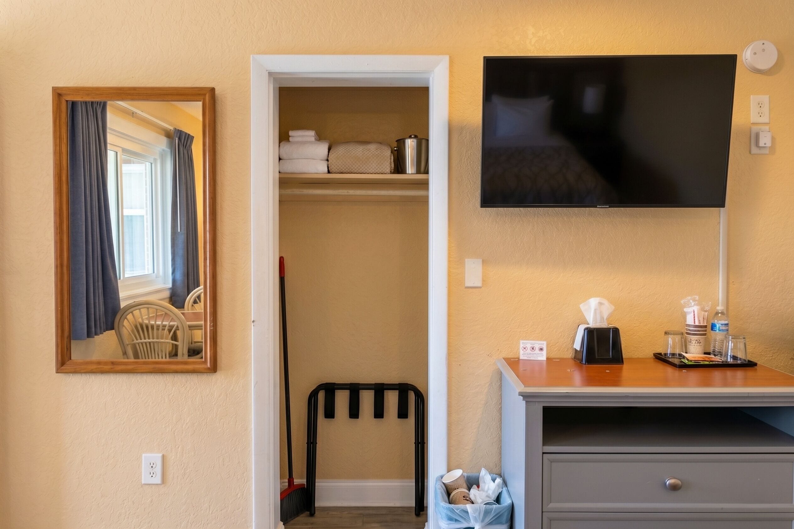 A hotel room with a TV, a desk with a tray, a trash bin, a coat rack, and a mirror.