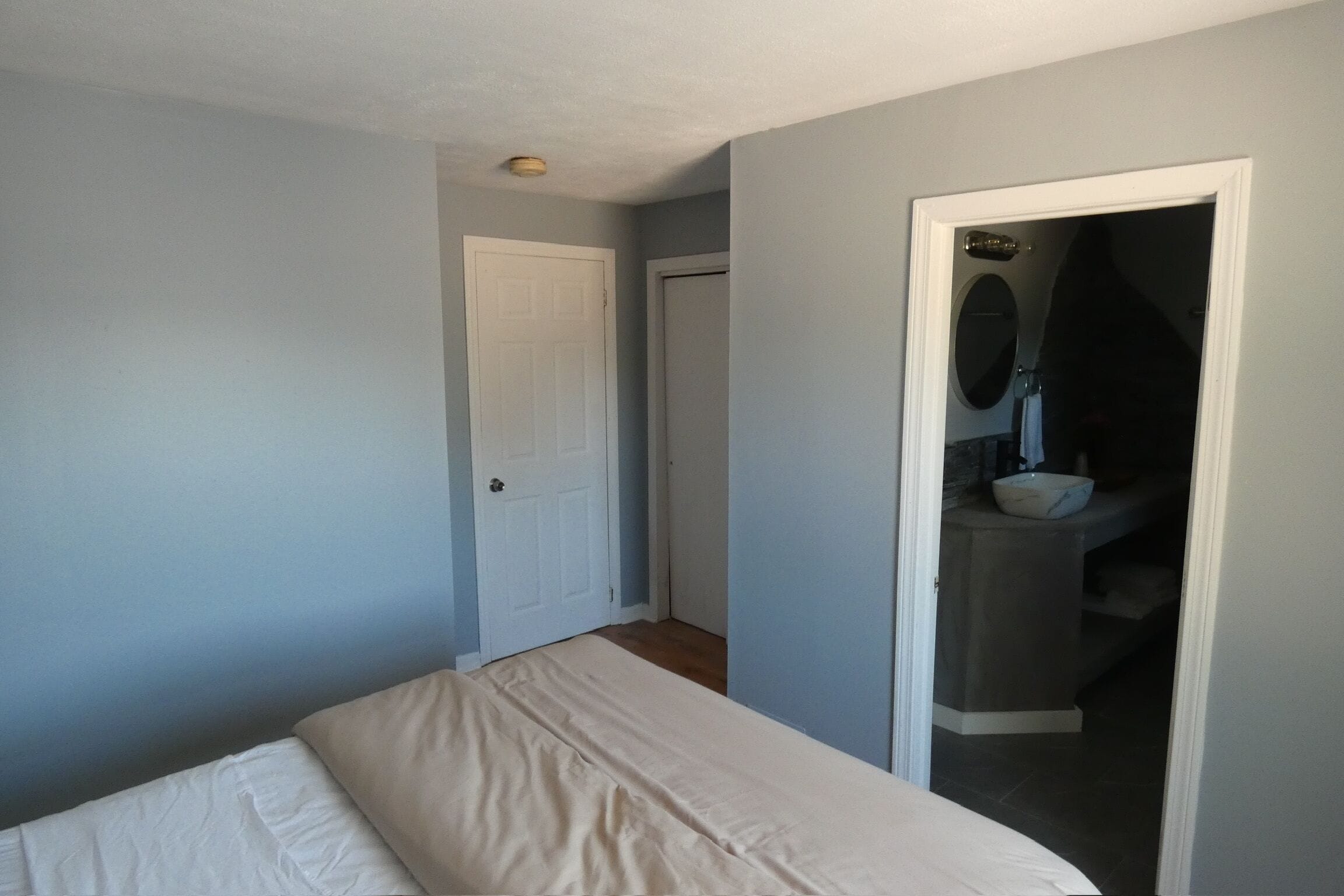 Un dormitorio con cama, una puerta y un baño visibles a través de un vano abierto.