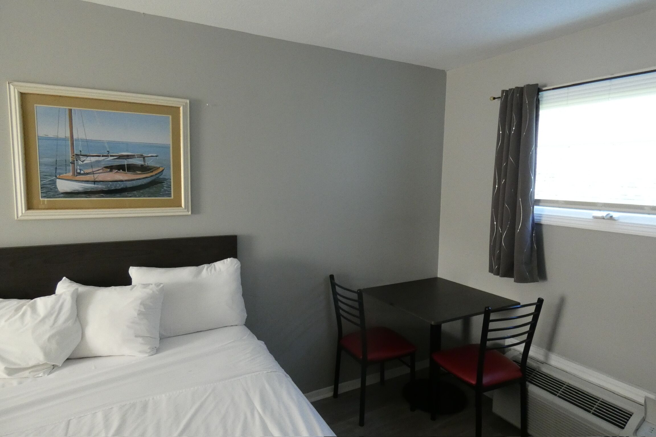 Una habitació d'hotel amb llit, escriptori, dues cadires, finestra amb cortines i un quadre al mur.