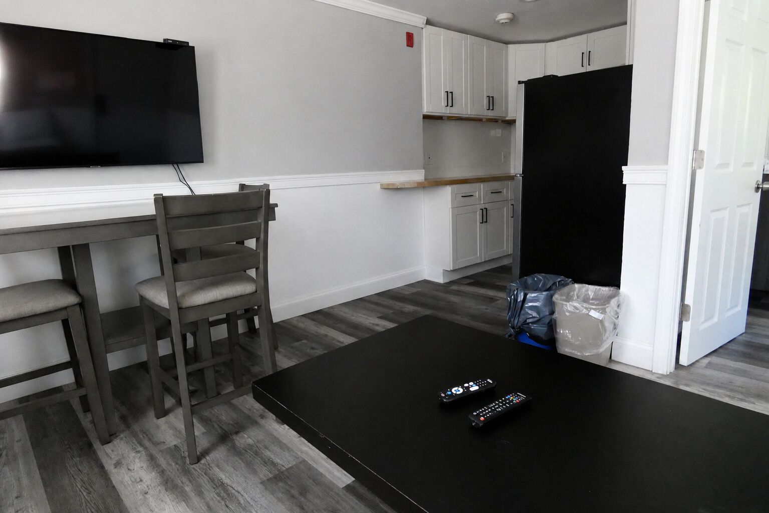 A room with a dining table, two chairs, a black coffee table, a TV mounted on the wall, and a kitchen area with cabinets and a refrigerator.