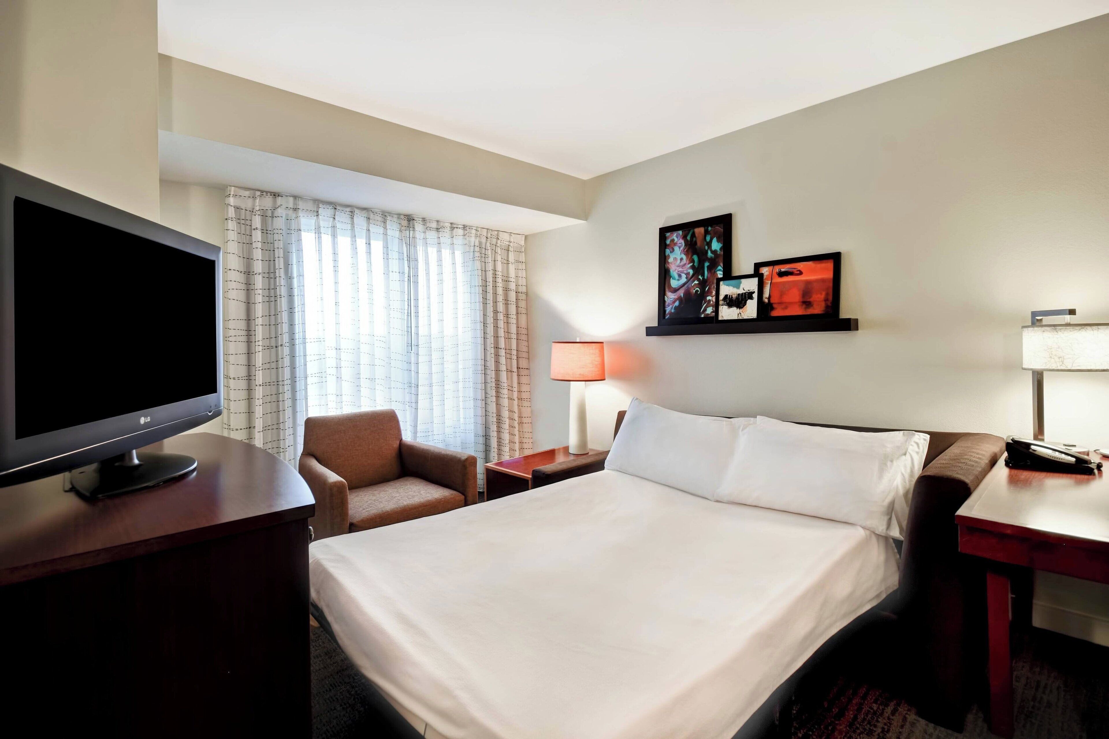 A hotel room with a bed, a TV, a desk, a chair, a lamp, and artwork on the wall.