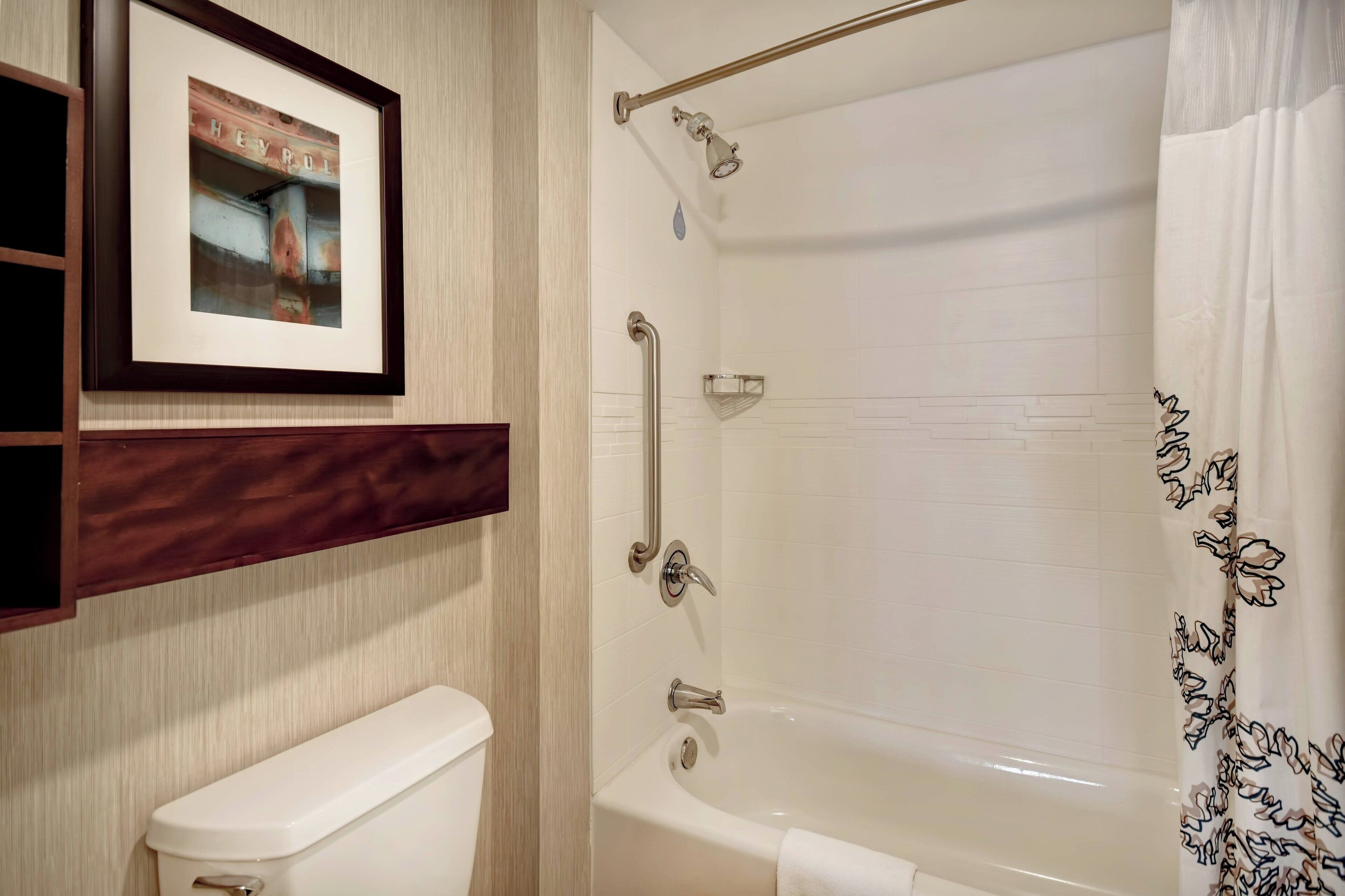 A bathroom with a white toilet, a framed picture on the wall, a bathtub with a shower curtain, and a handrail.