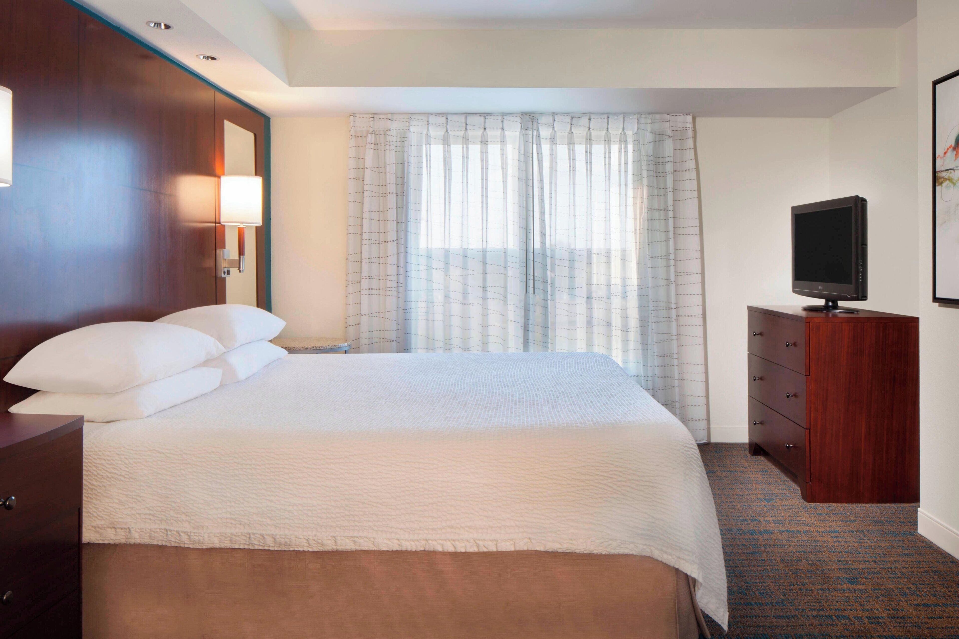 A hotel room with a large bed, a wooden dresser, a television, and a window with curtains.