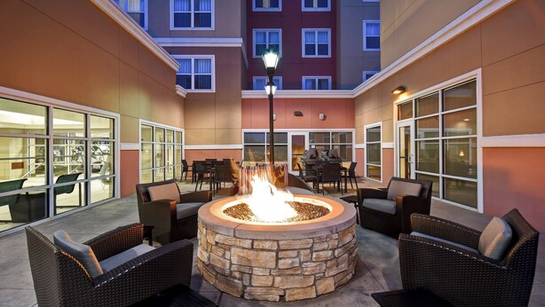 An outdoor patio with a stone fire pit surrounded by wicker furniture, large windows, and a building in the background. - Residence Inn by Marriott Stillwater (Stillwater)