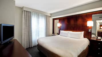 Habitación de hotel con una cama grande, televisión de pantalla plana, mesita de noche de madera, espejo y ventana con cortinas.