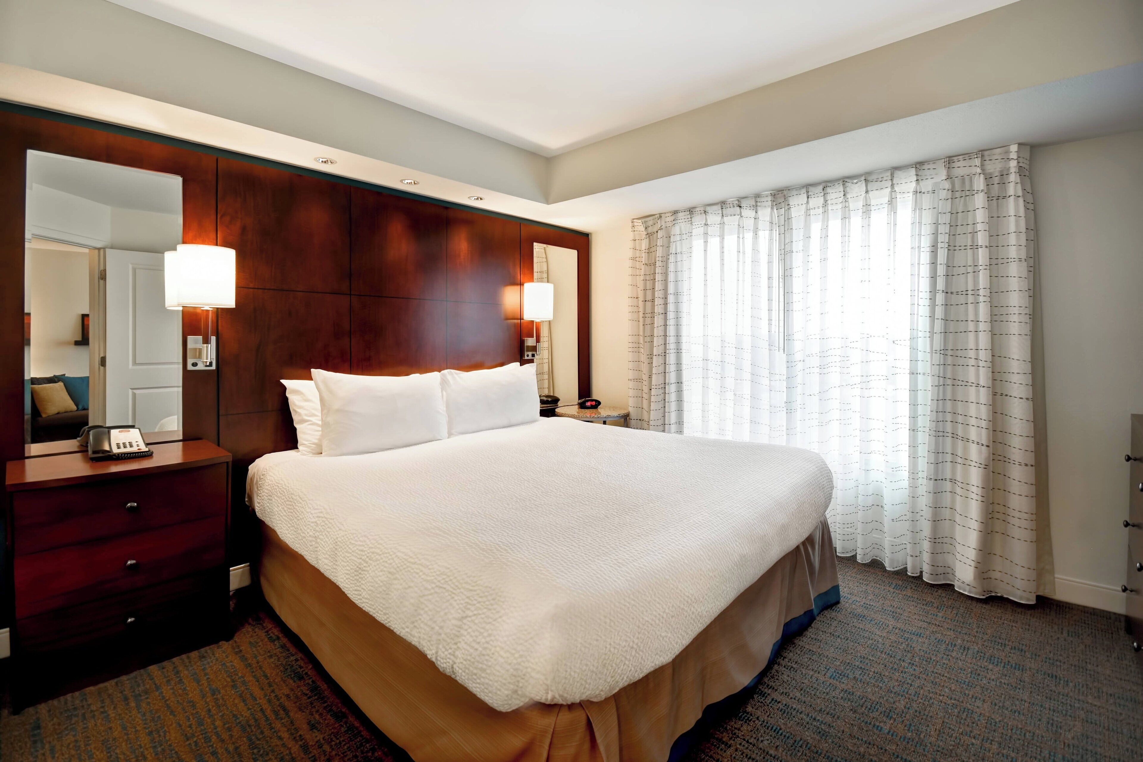 A hotel room with a large bed, a wooden headboard, a bedside table with a lamp, a mirror, and a window with curtains.
