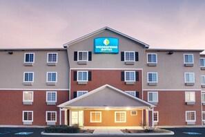 Un hotel de varios pisos con un llamativo letrero de "Woodspring Suites". Presenta fachada de ladrillo, múltiples ventanas y una entrada central.