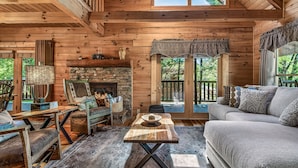 A cozy cabin interior with a stone fireplace, wooden furniture, and large windows offering a view of the outdoors.
